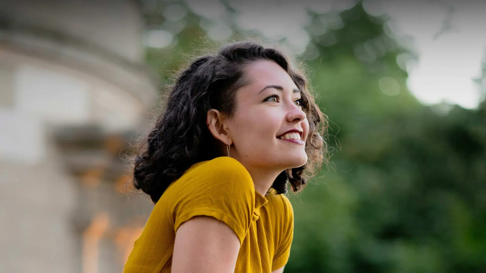 Smiling woman in a yellow shirt, outdoors with green foliage in the background.