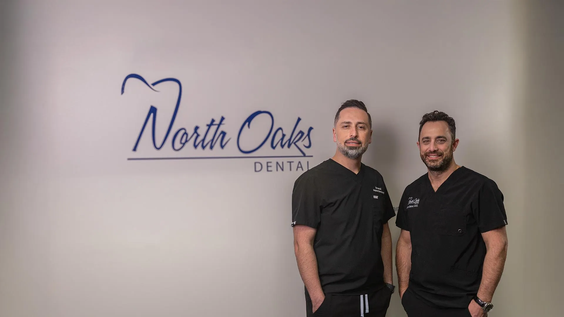 Two male dentists in black scrubs standing in front of North Oaks Dental logo on a white wall