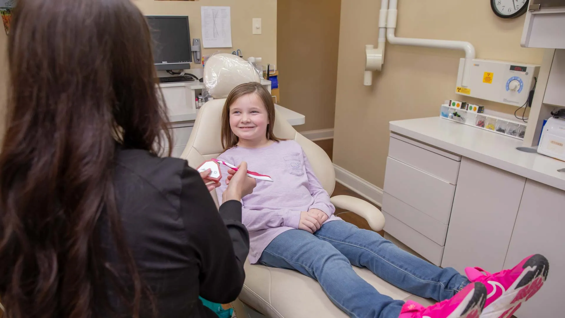 Young girl smiles while dentist explains dental care using a toothbrush model in a modern dental office.