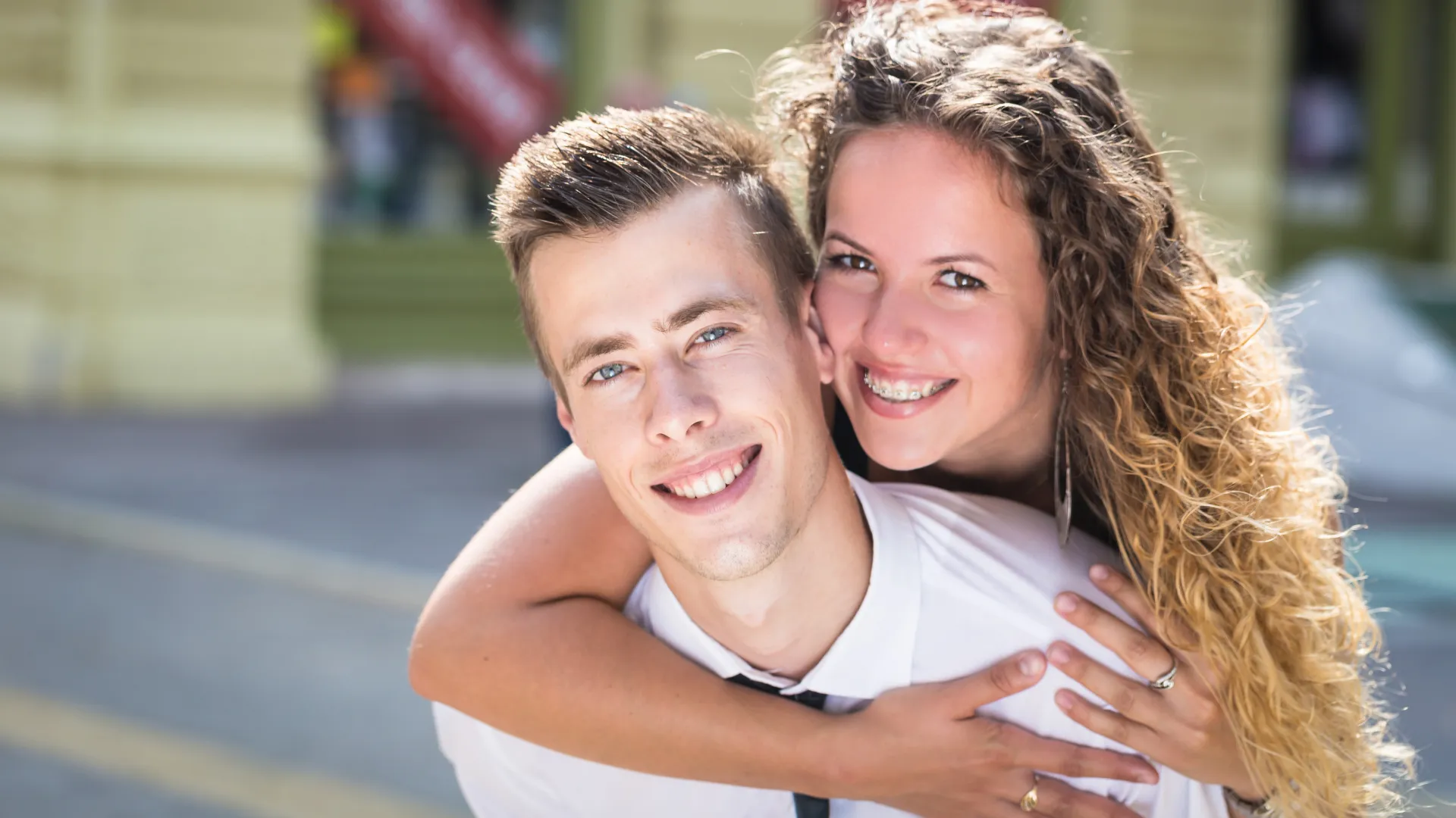 Smiling young couple with woman hugging man from behind with six month smiles braces outdoors in an urban setting.