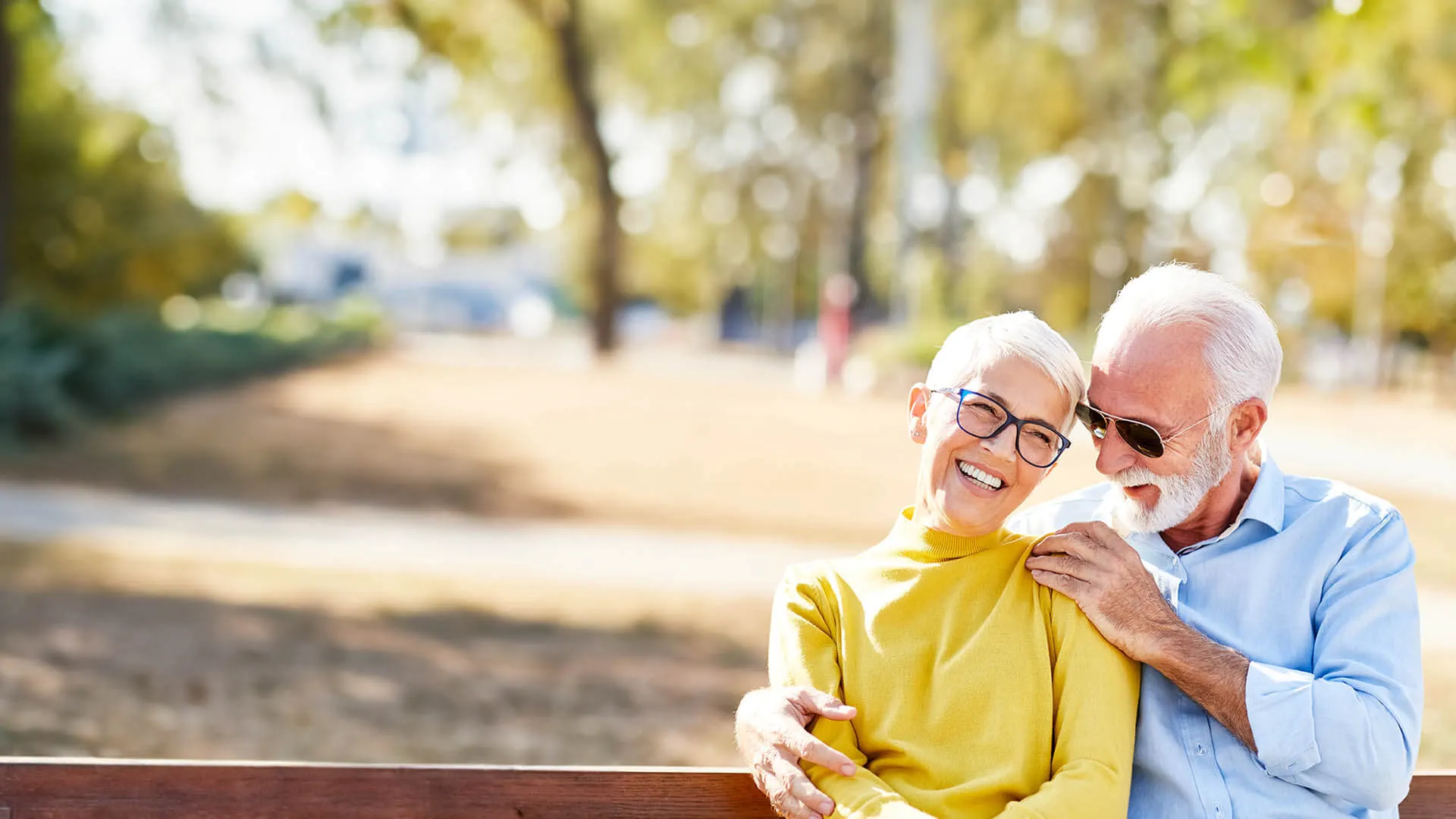 Happy senior couple sitting on a park bench enjoying a sunny day with trees in the background