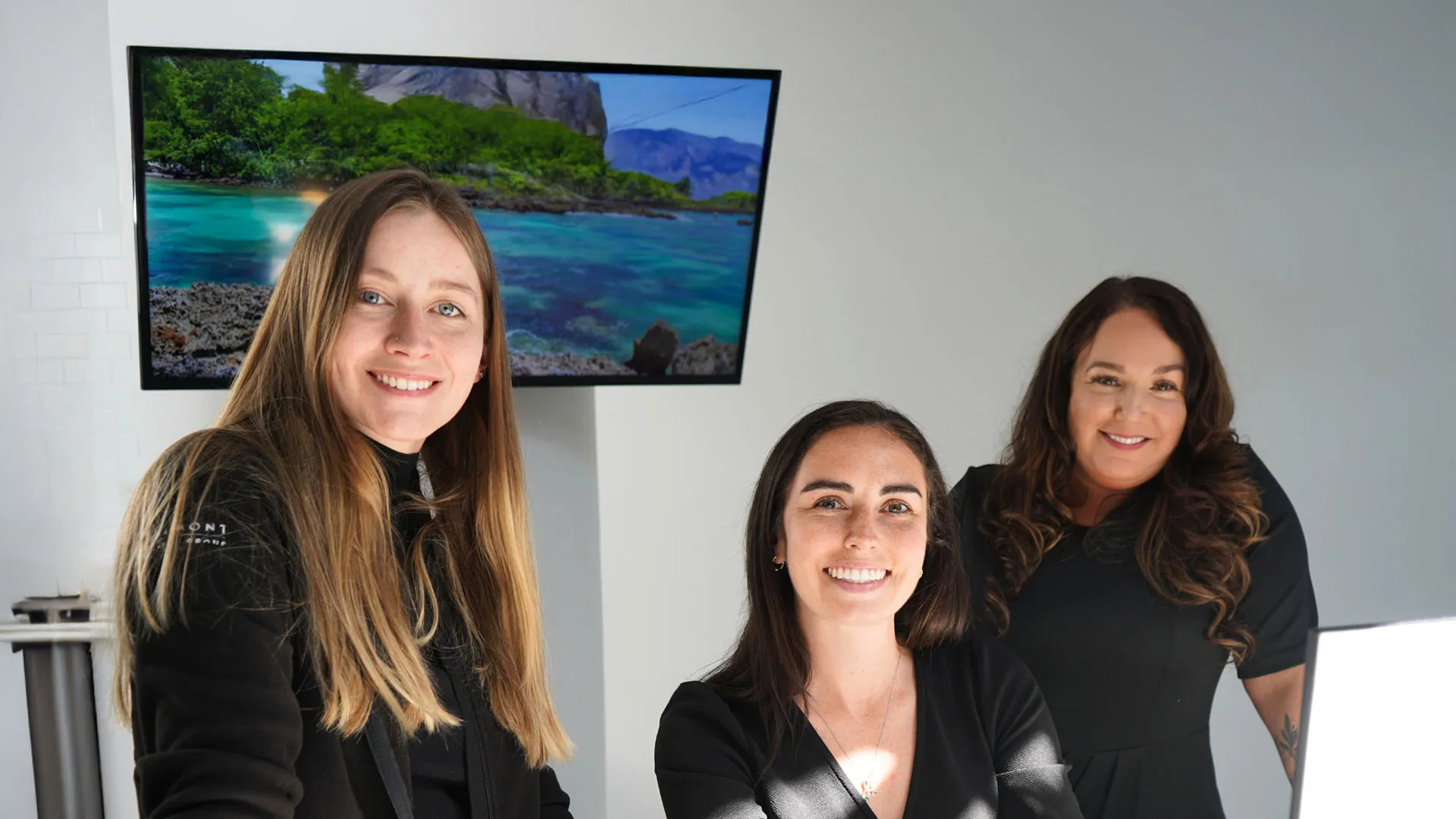 Three women smiling in a bright space with a nature scene displayed on a screen behind them.
