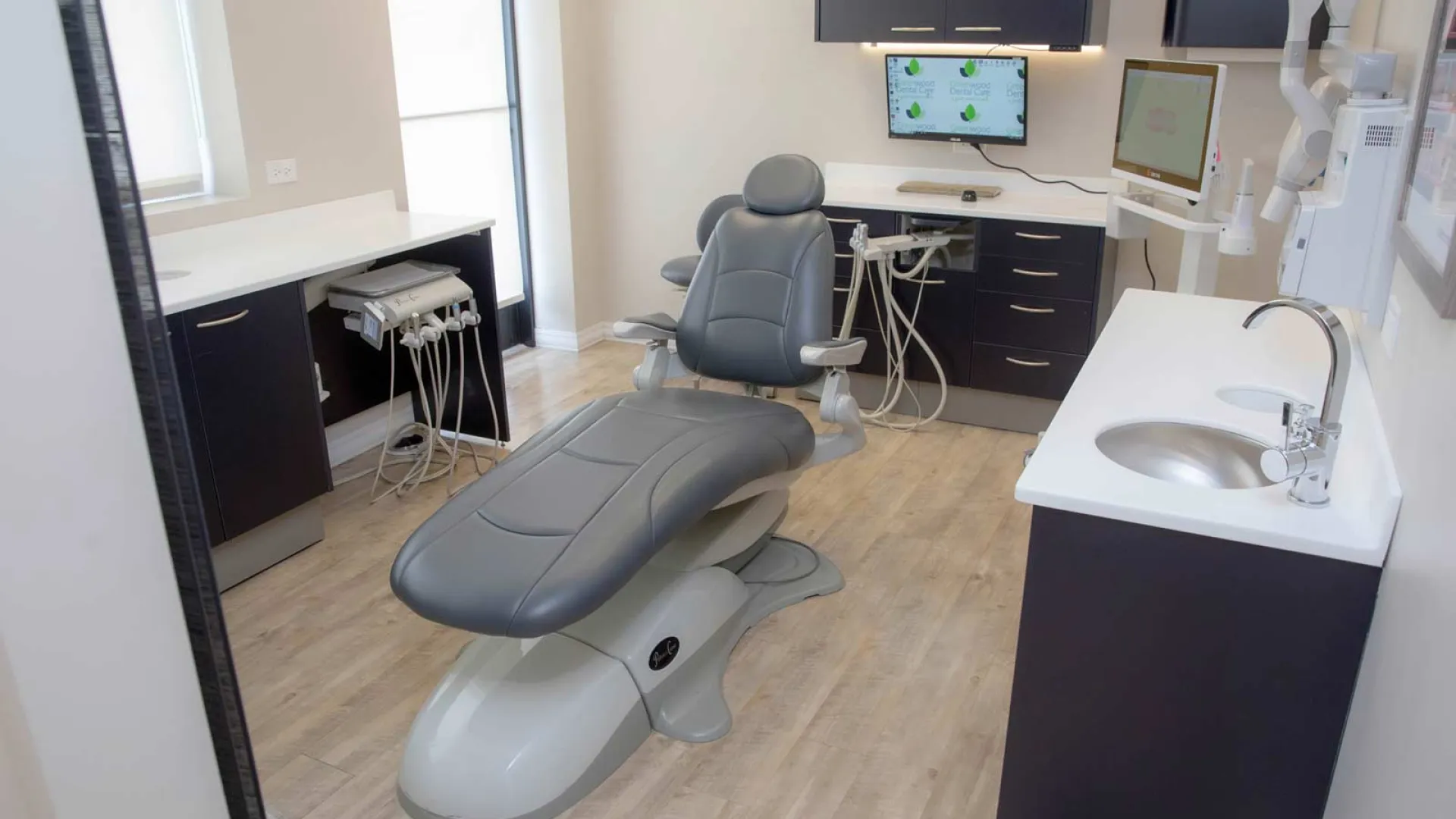 Modern dental clinic room with adjustable chair, dental tools, sink, and computer screens on wooden floor.