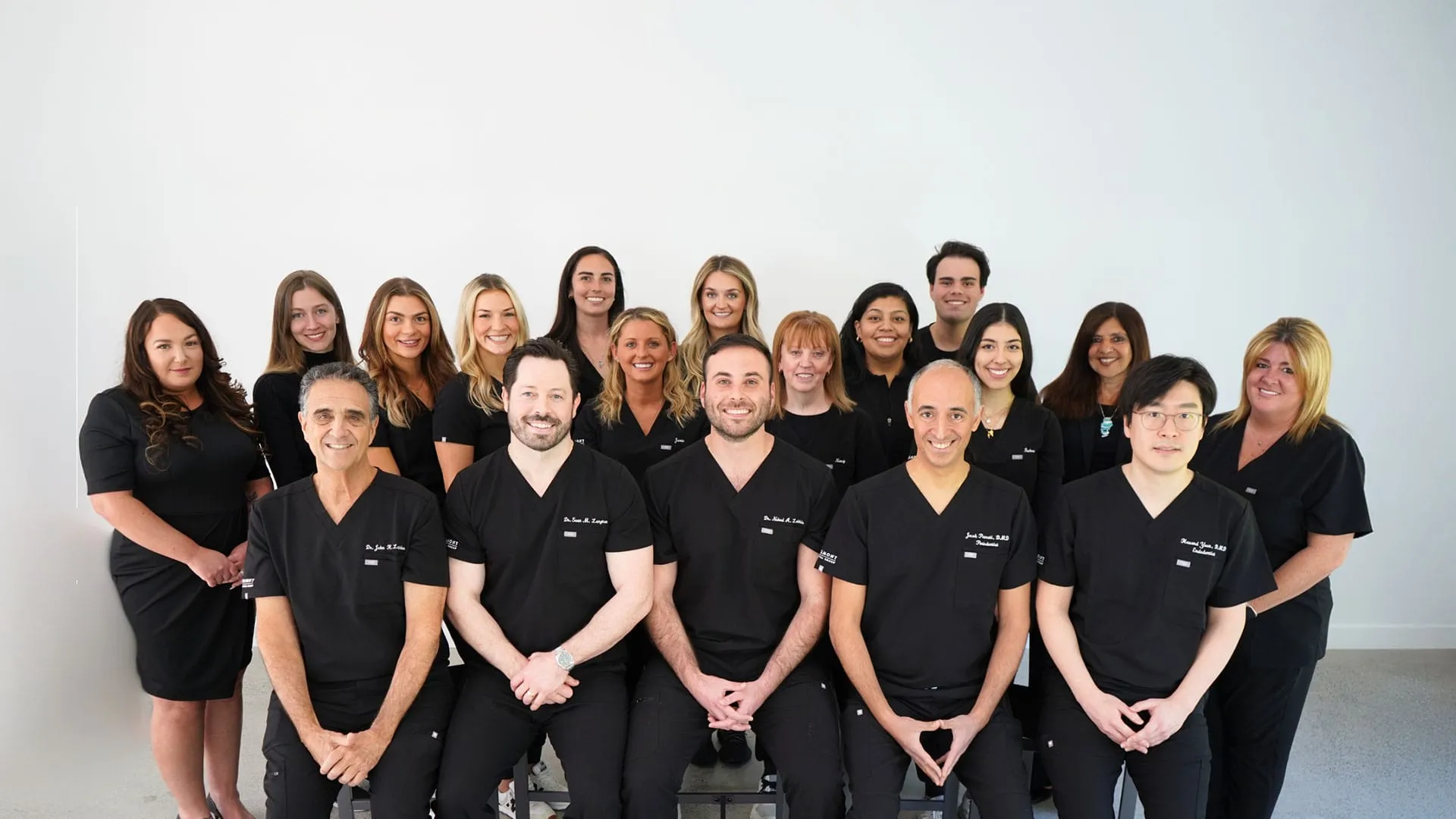 Group portrait of medical professionals wearing black scrubs and formal attire standing and sitting against a white background