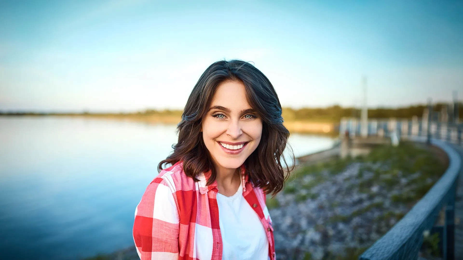 Smiling young woman with blue eyes and brown hair in red plaid shirt standing near a lake on a clear day