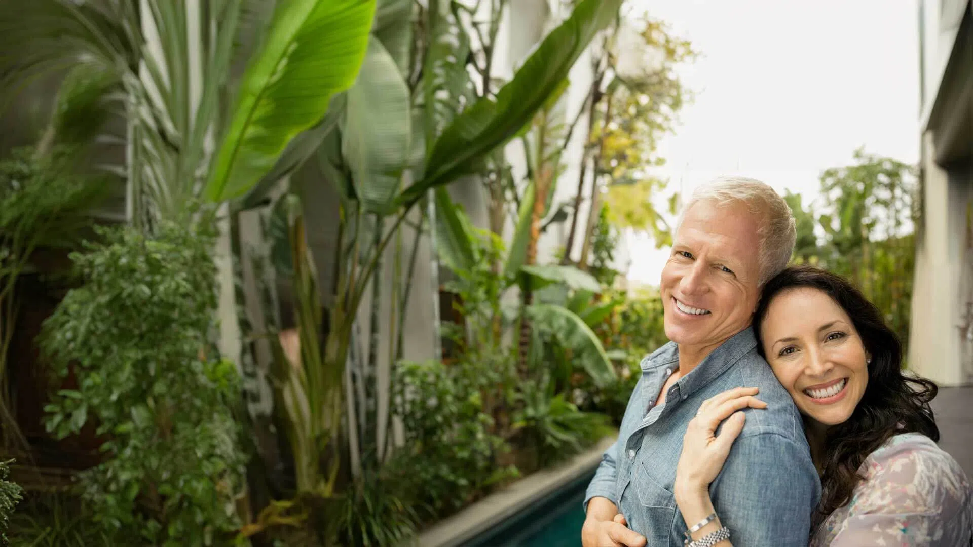 Smiling mature couple embraces outdoors near lush green plants and a narrow pool on a sunny day