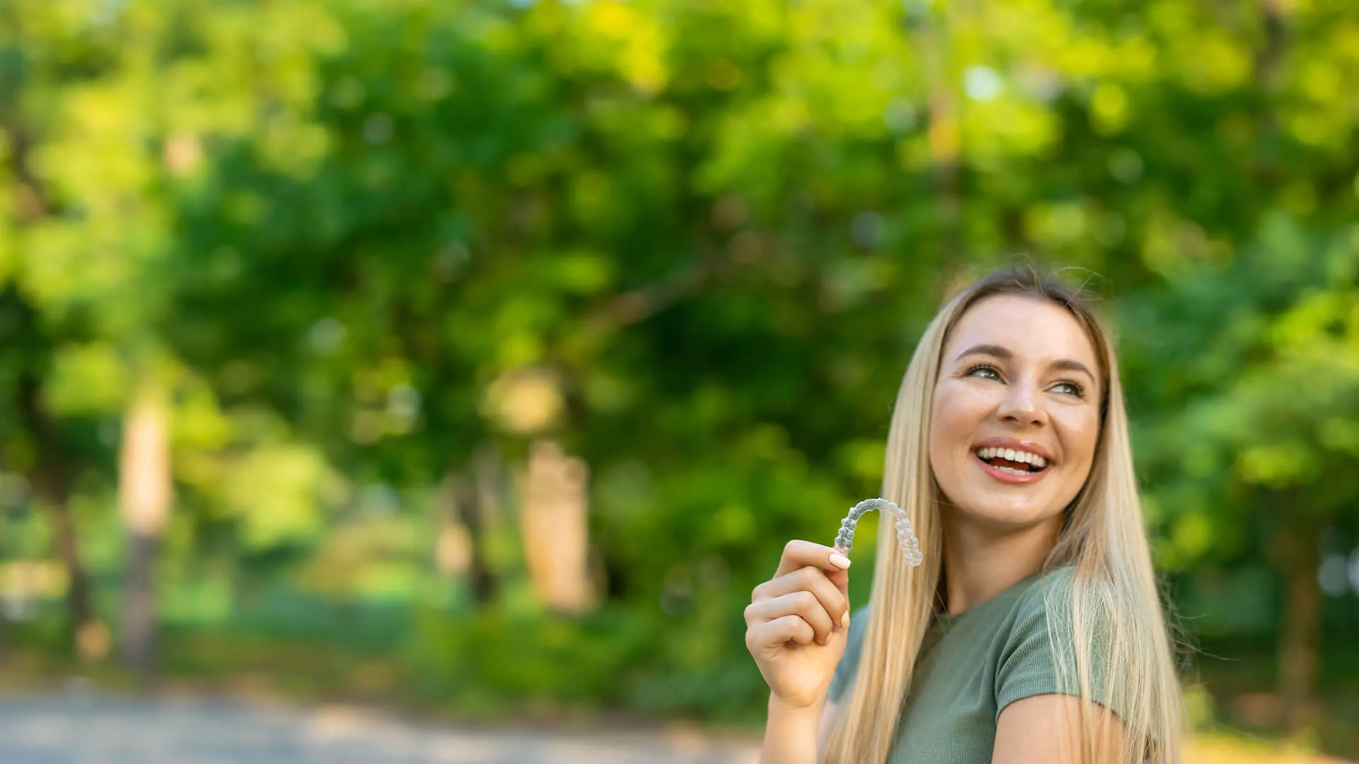 Smiling woman outdoors holding an invisible aligner with greenery in the background.
