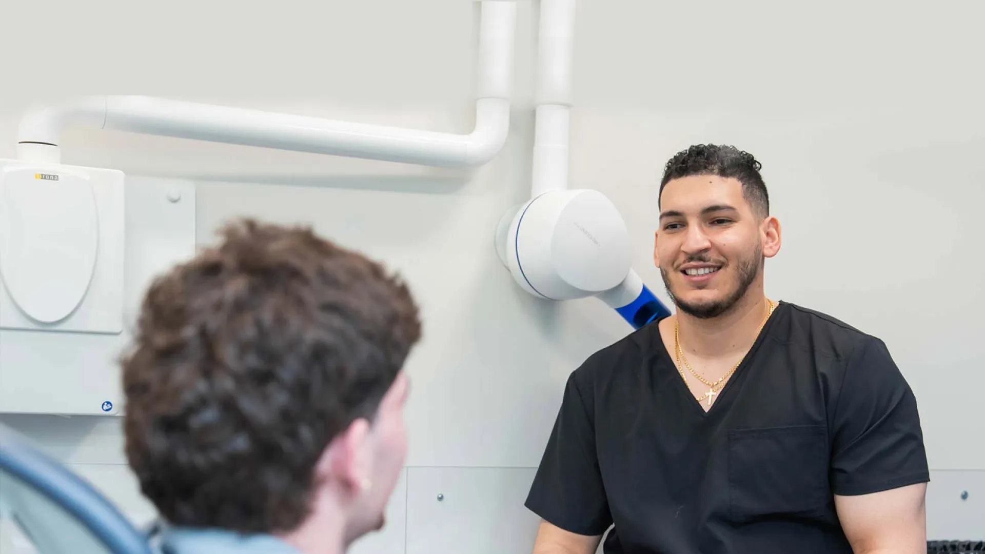 Dentist in black scrubs talking to patient in a modern dental clinic with white walls and equipment.