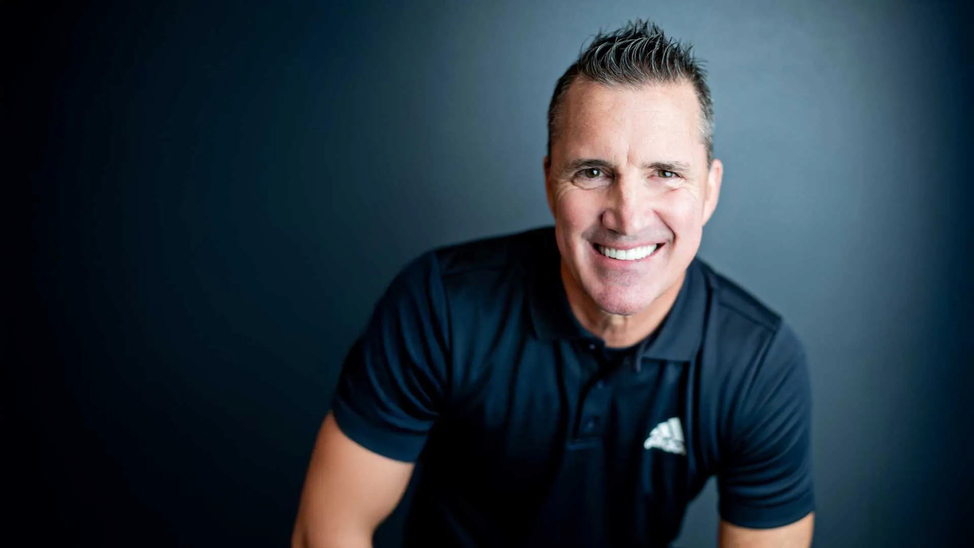 Smiling middle-aged man with short hair wearing a black Adidas polo shirt against a dark background