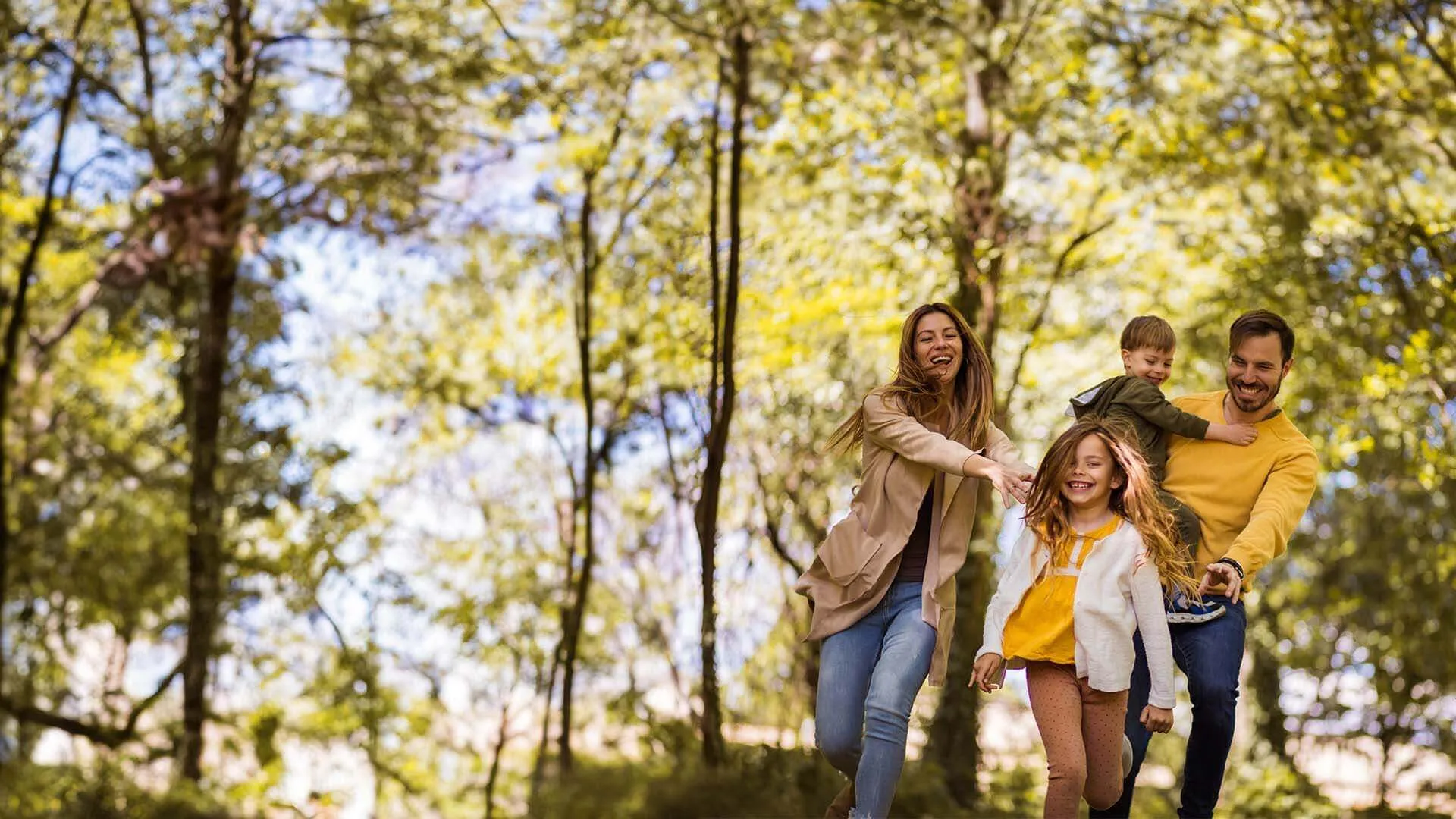 Happy family of four walking together in a sunny forest with green trees during autumn.