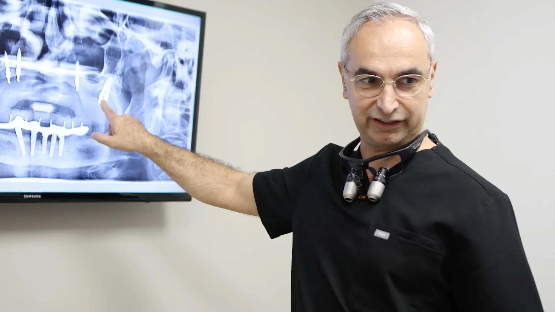 Dentist in black scrubs pointing at dental X-ray on screen with dental loupes around neck in clinic.