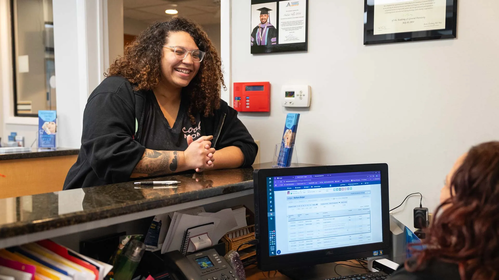 Smiling woman with curly hair talks to receptionist at office counter with computer and phone.