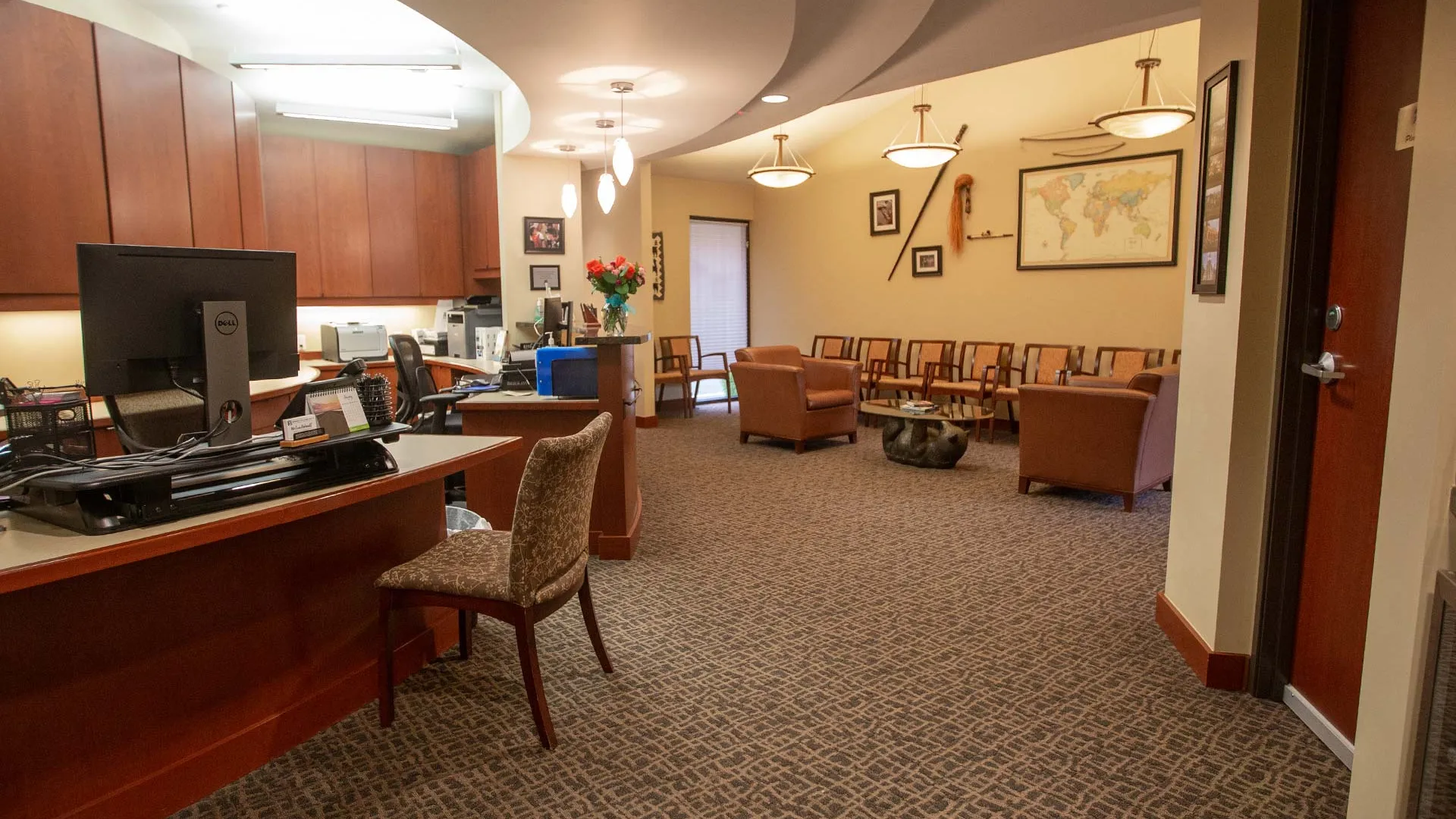 Spacious waiting area with wooden reception desk, computer, comfortable chairs, and wall decorations in warm lighting.