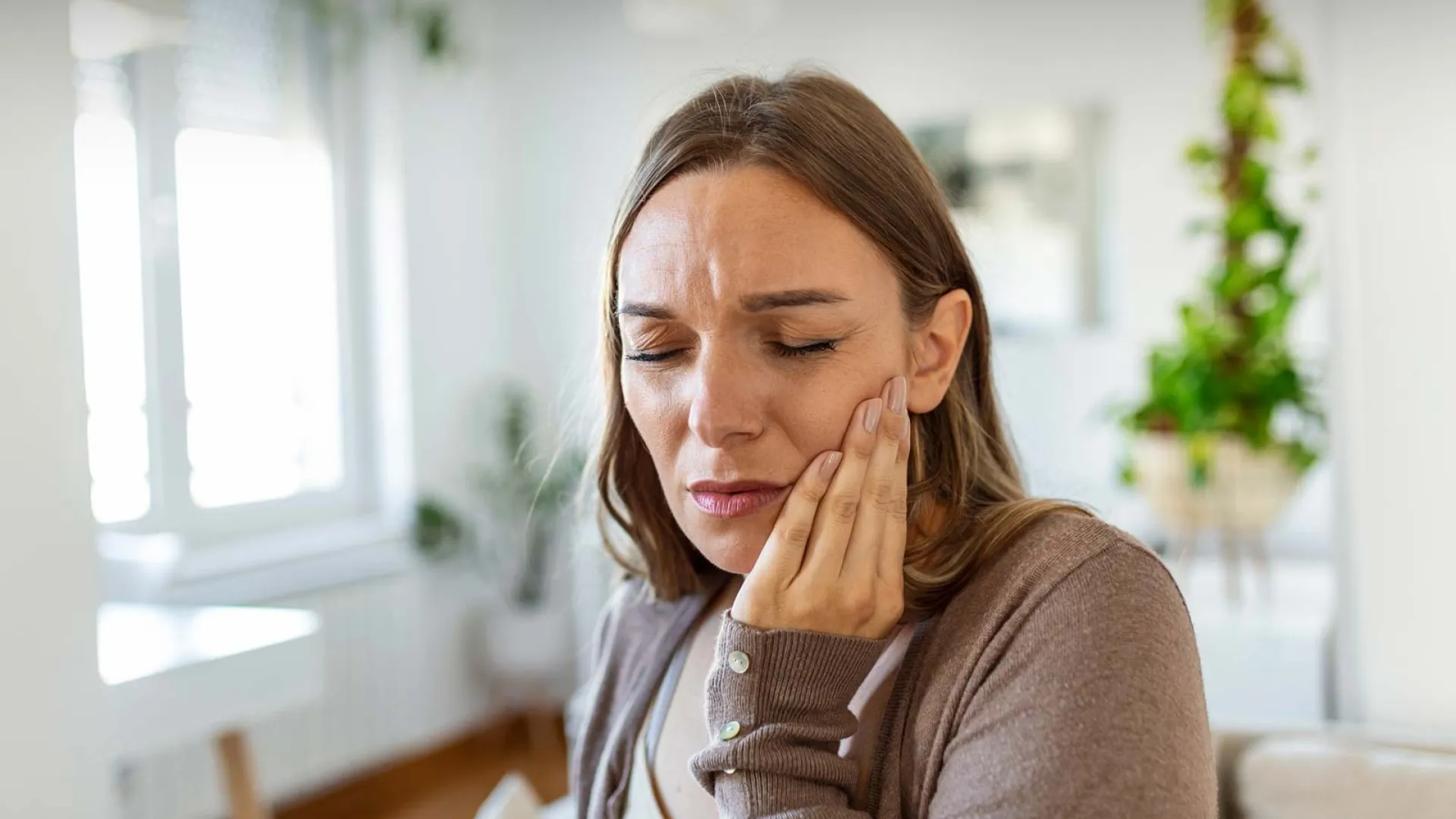 Woman experiencing dental pain while sitting in a modern, plant-filled living room.