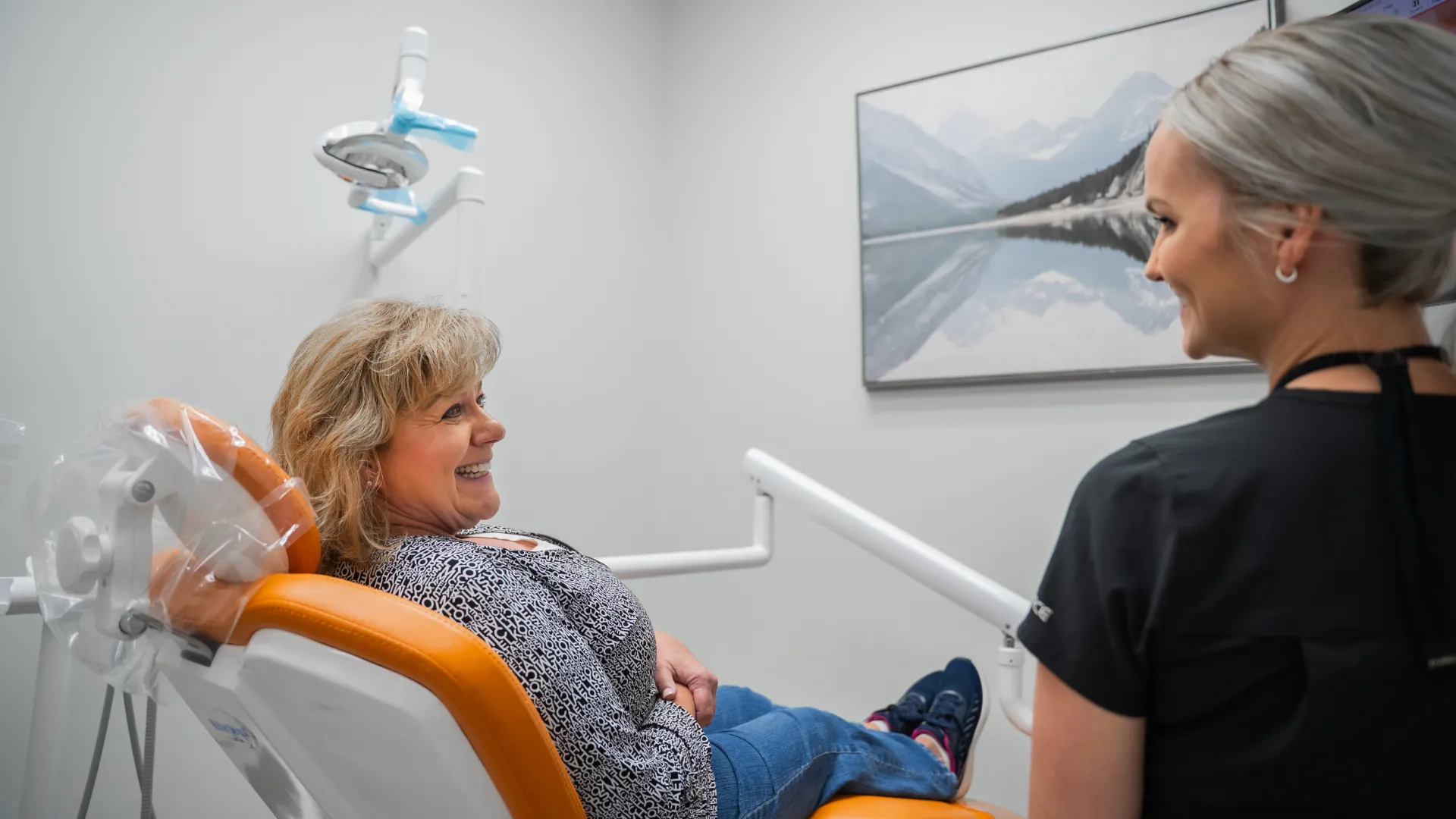 Smiling female patient sits in dental chair talking to female dentist in clinic with mountain artwork