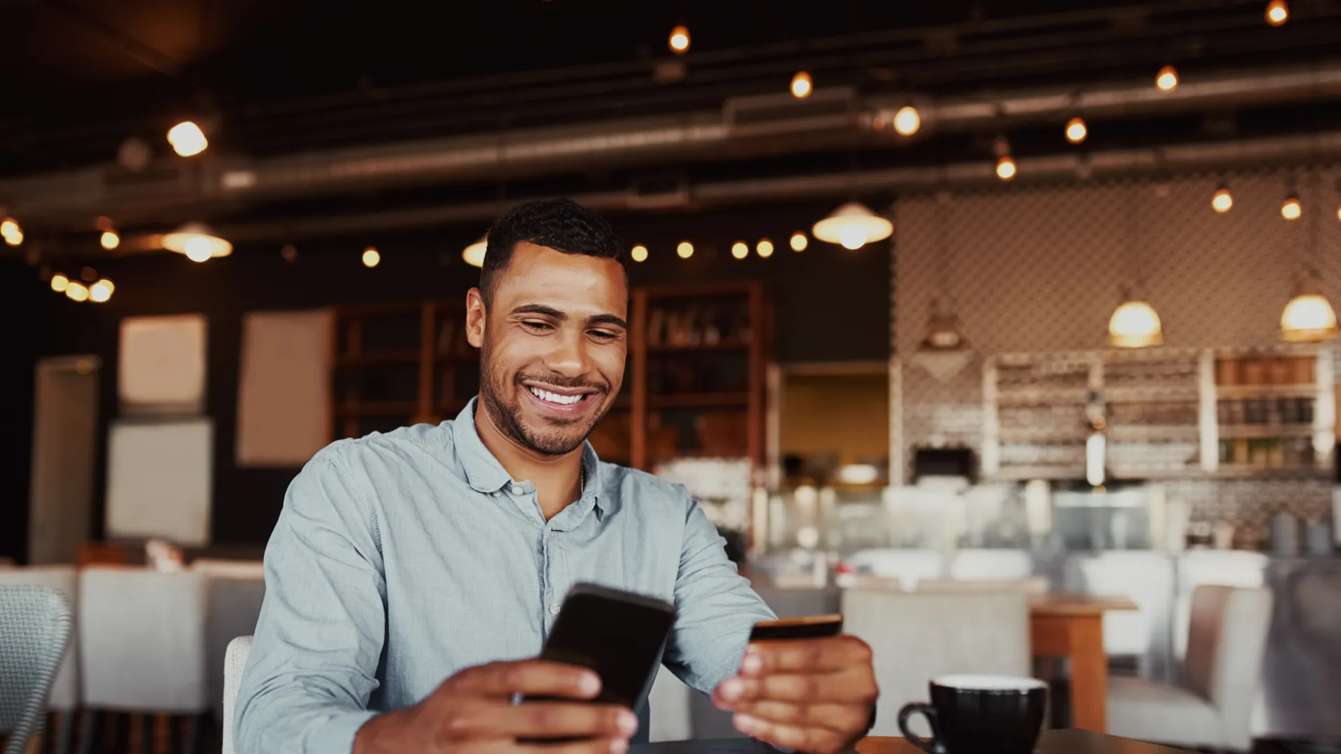 Smiling man scanning credit card with smartphone in cozy cafe with coffee and notebook on table.