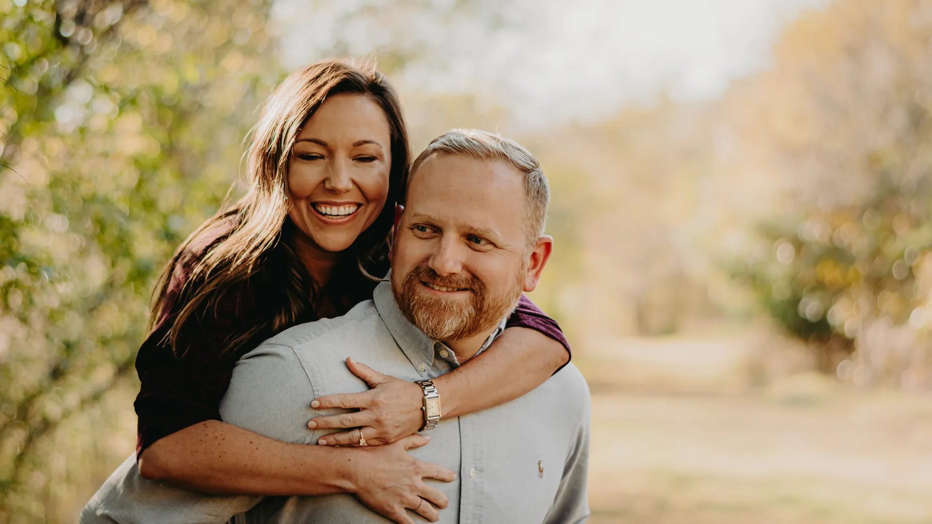 Smiling woman hugs man from behind outdoors in a bright, natural setting with fall foliage in background