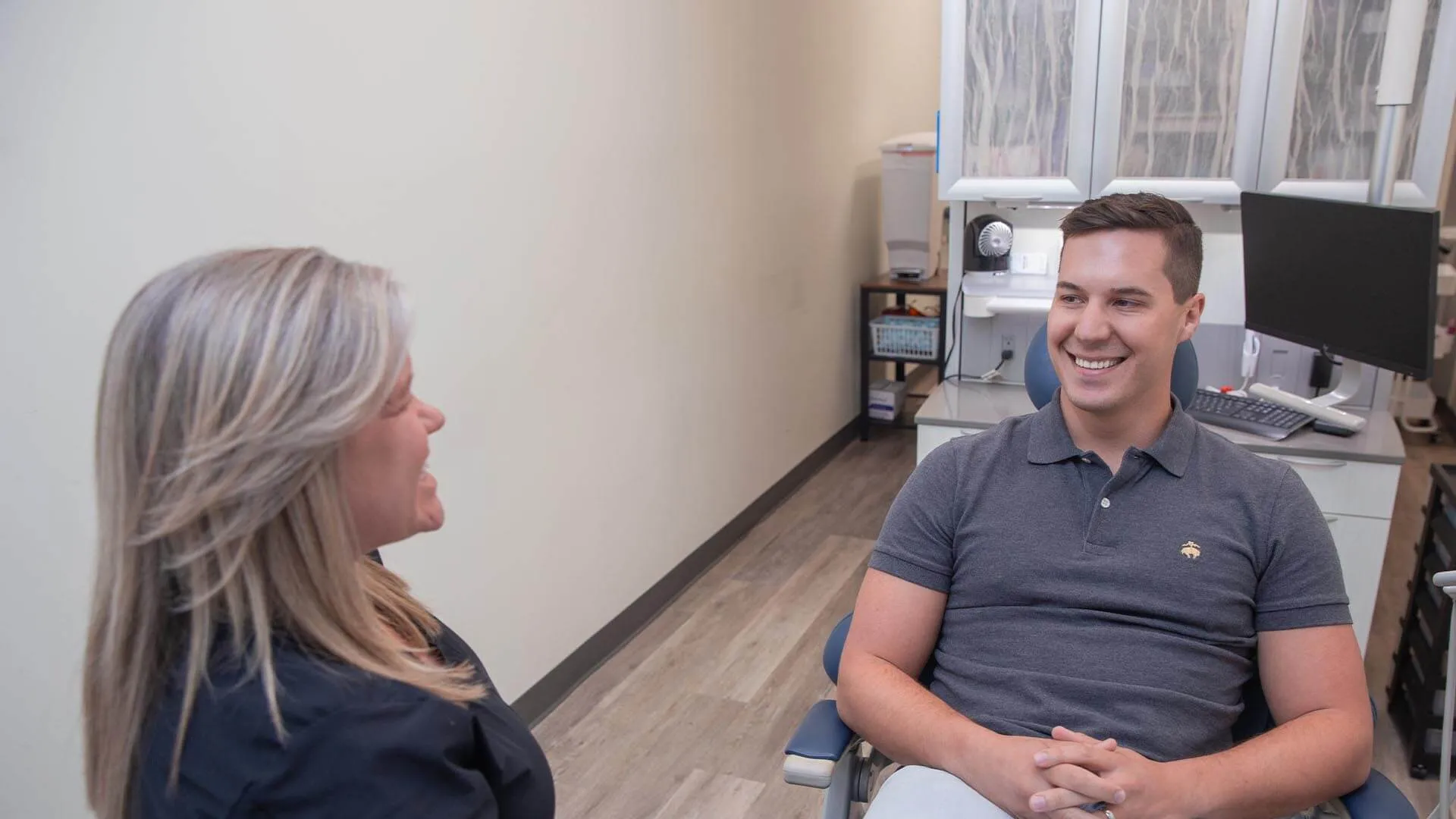 Smiling man and woman having a friendly conversation in a modern office setting with computer and shelves.