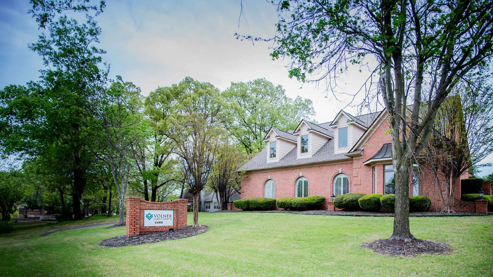 Brick office building with green lawn and trees under a cloudy blue sky during springtime.