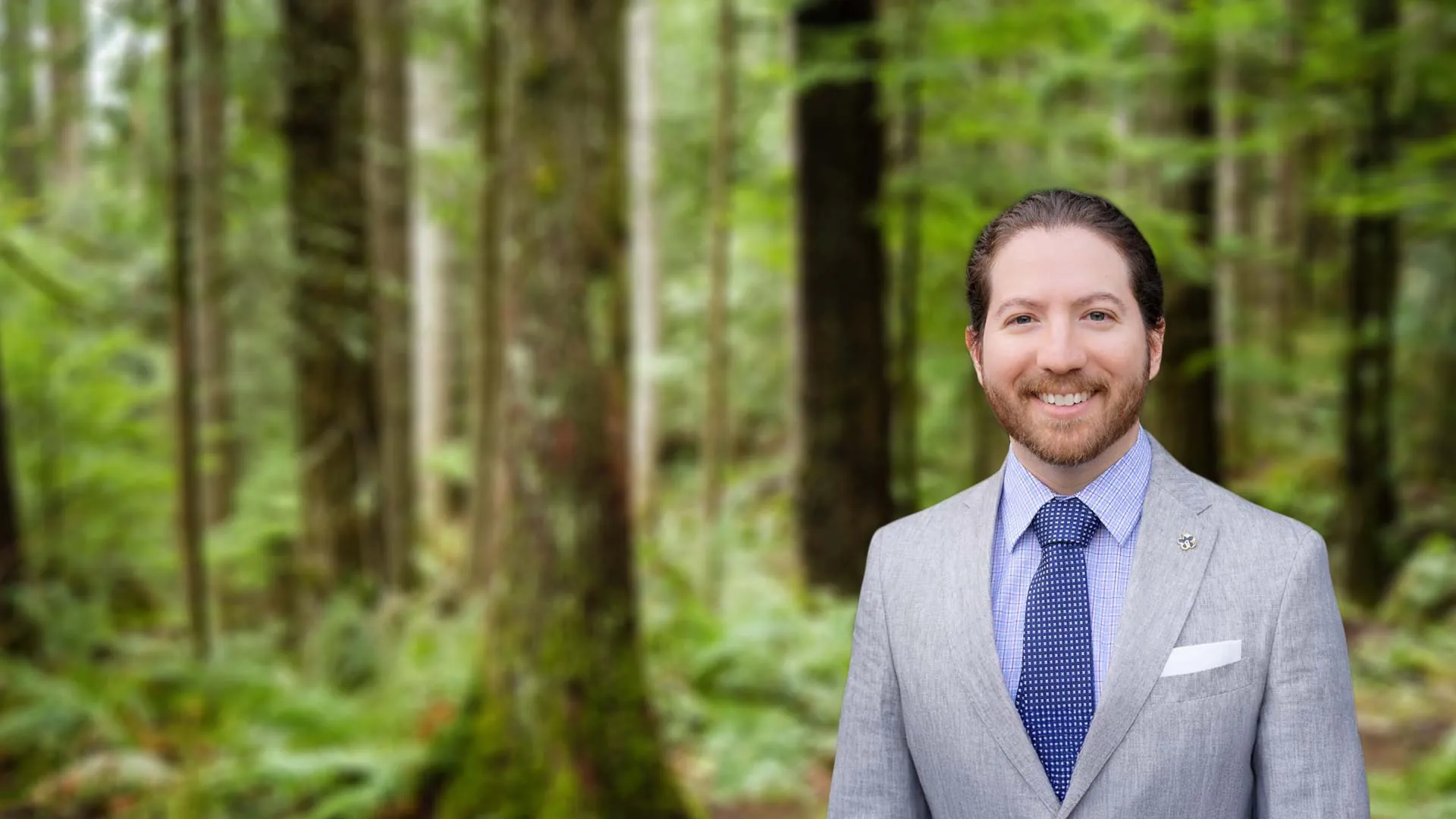 Smiling man in light gray suit and blue tie standing in a lush green forest with tall trees.