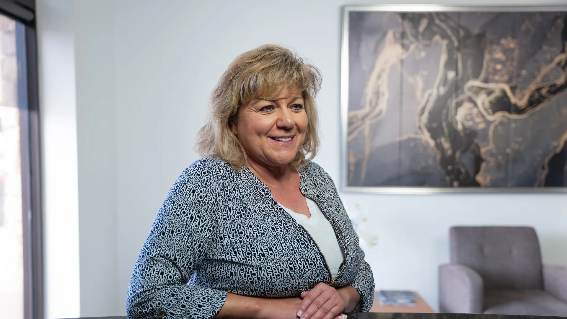 Smiling middle-aged woman in patterned blouse sitting in modern office with abstract wall art and gray chair.