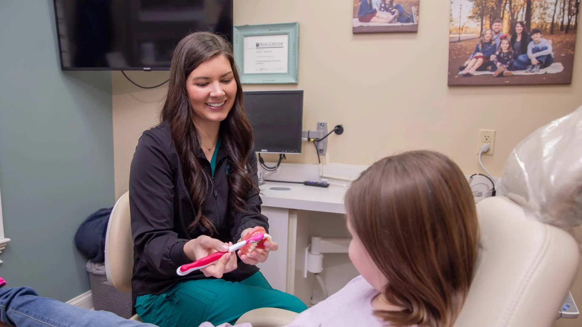 Dentist explains proper brushing technique to young girl in a dental office with family photo in background