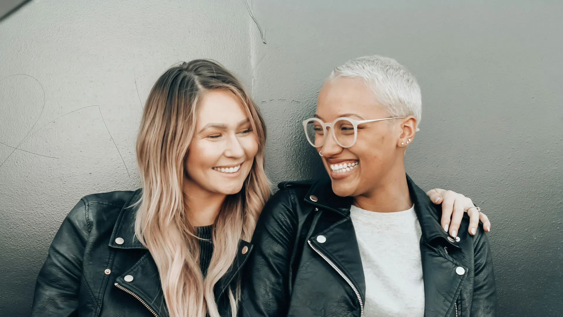 Two women in leather jackets smiling and embracing each other against a gray wall background.