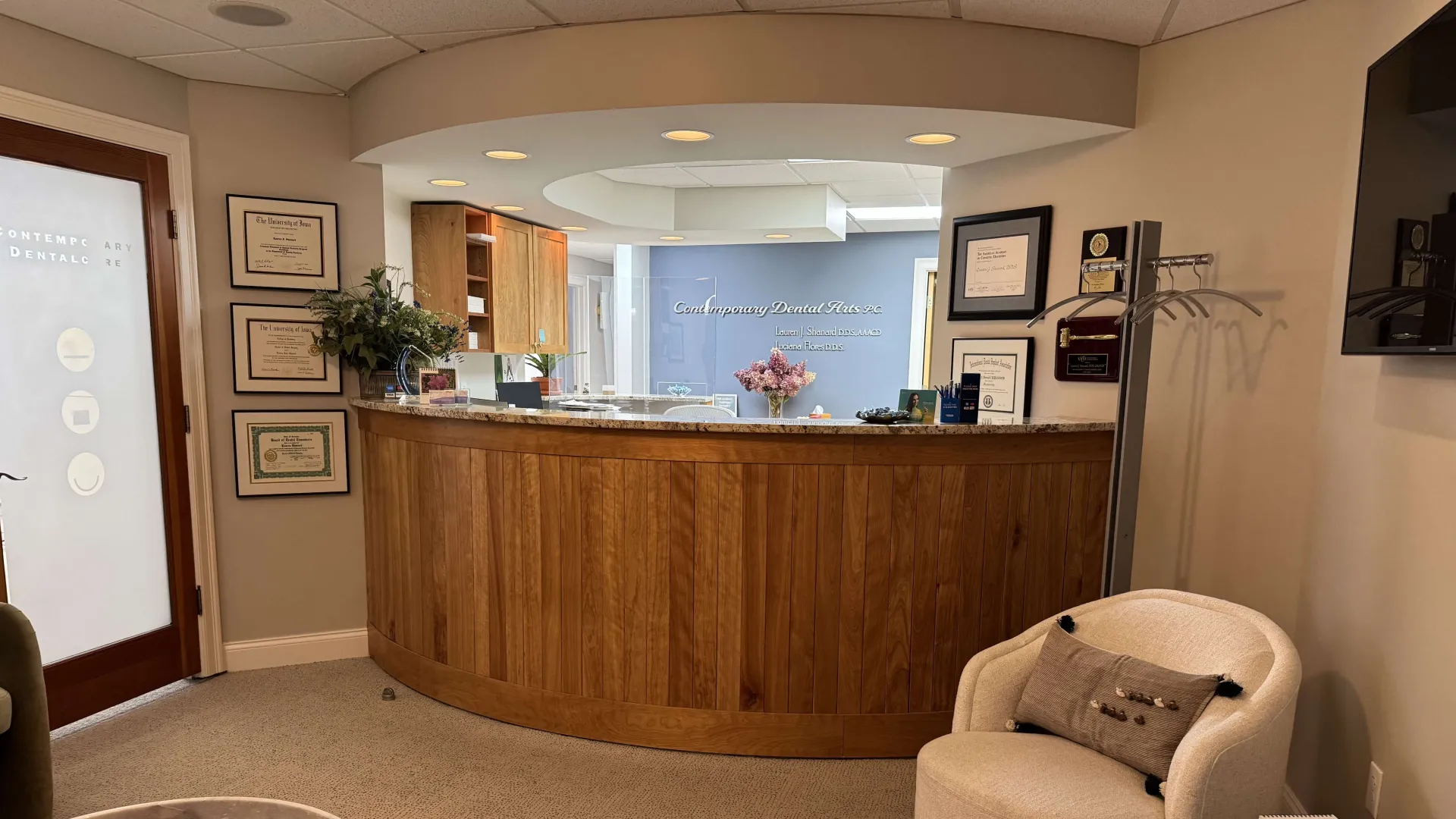 Reception area of a dental office with wooden desk, certificates on walls, chair, and tv screen.