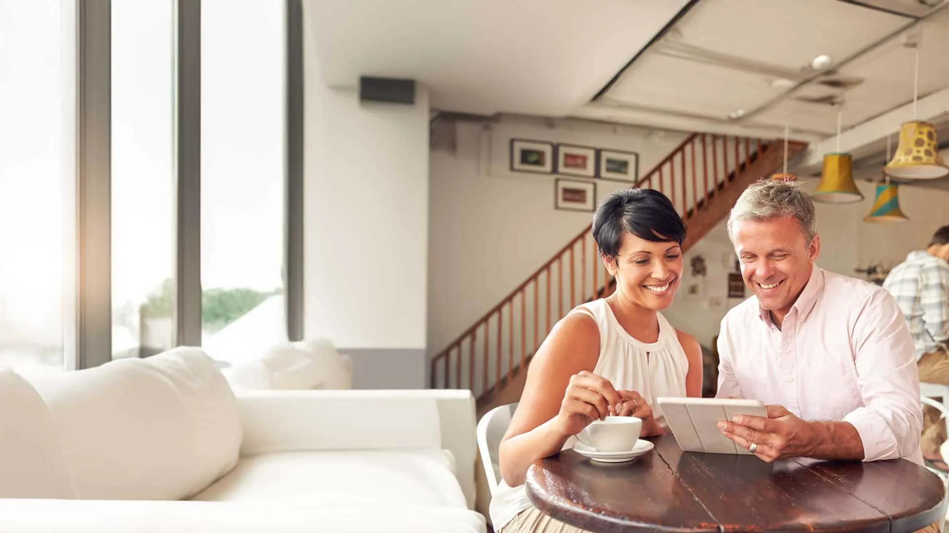 Smiling couple sitting at a wooden table in a bright cafe looking at a tablet and drinking coffee together