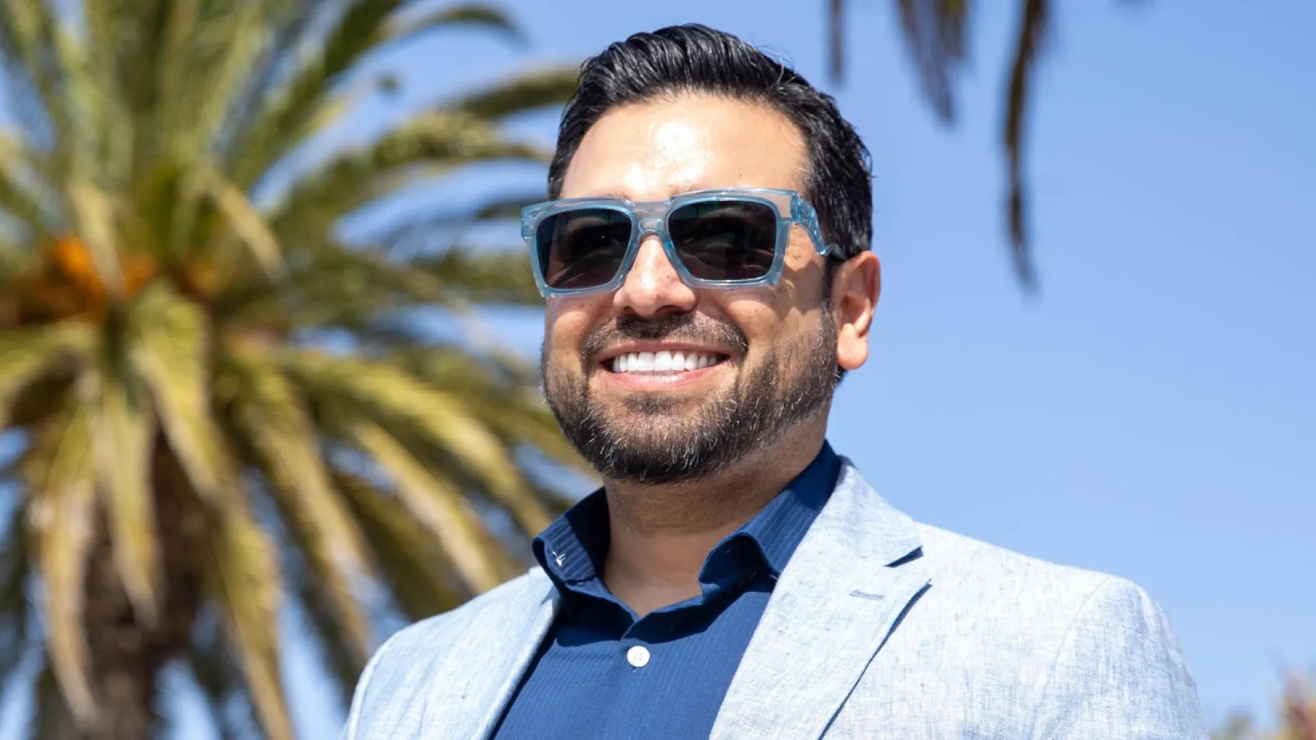Man smiling in sunglasses against a backdrop of palm trees and blue sky.