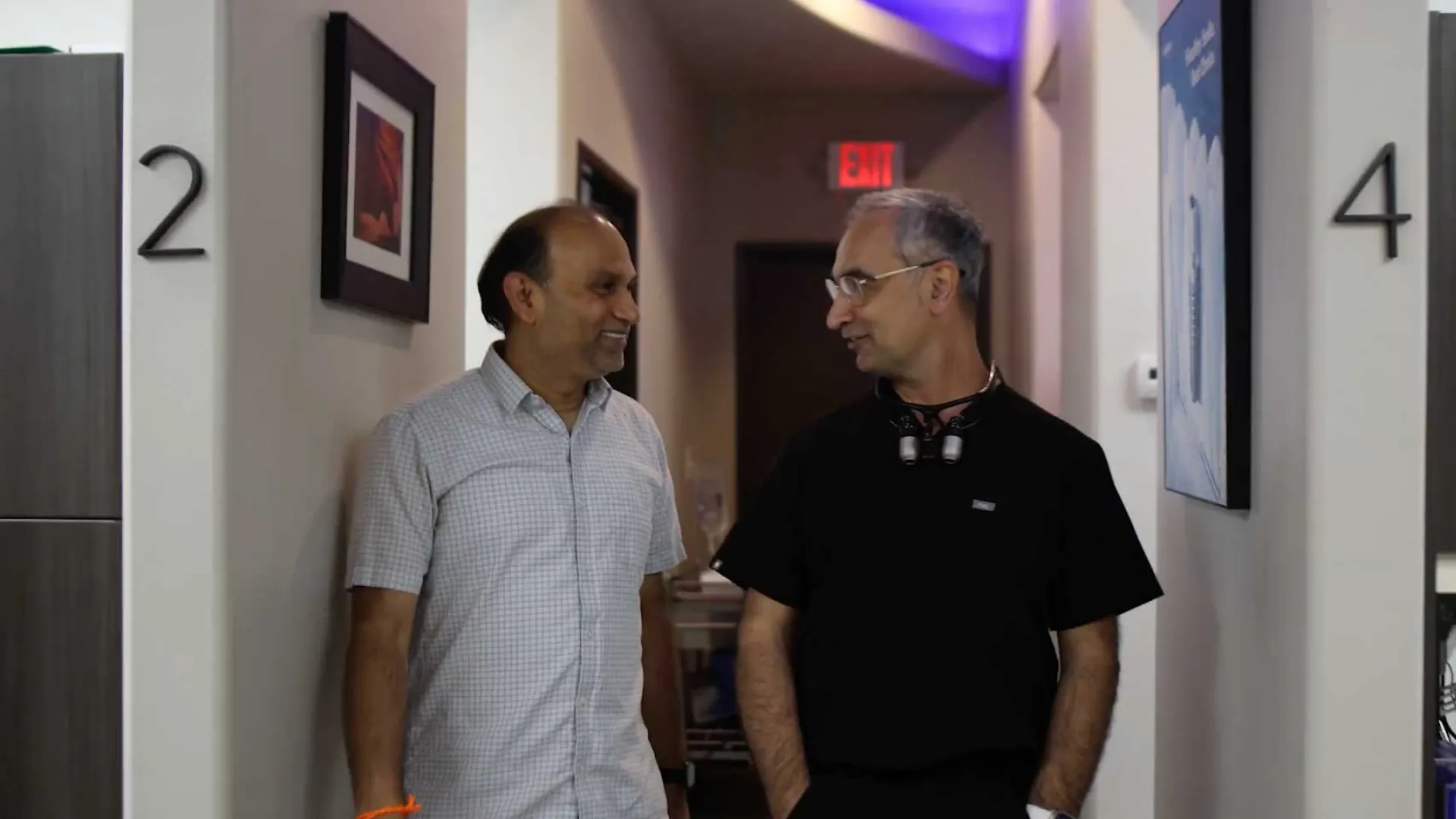Two men walking and talking in a hallway between rooms numbered 2 and 4 with exit sign in background