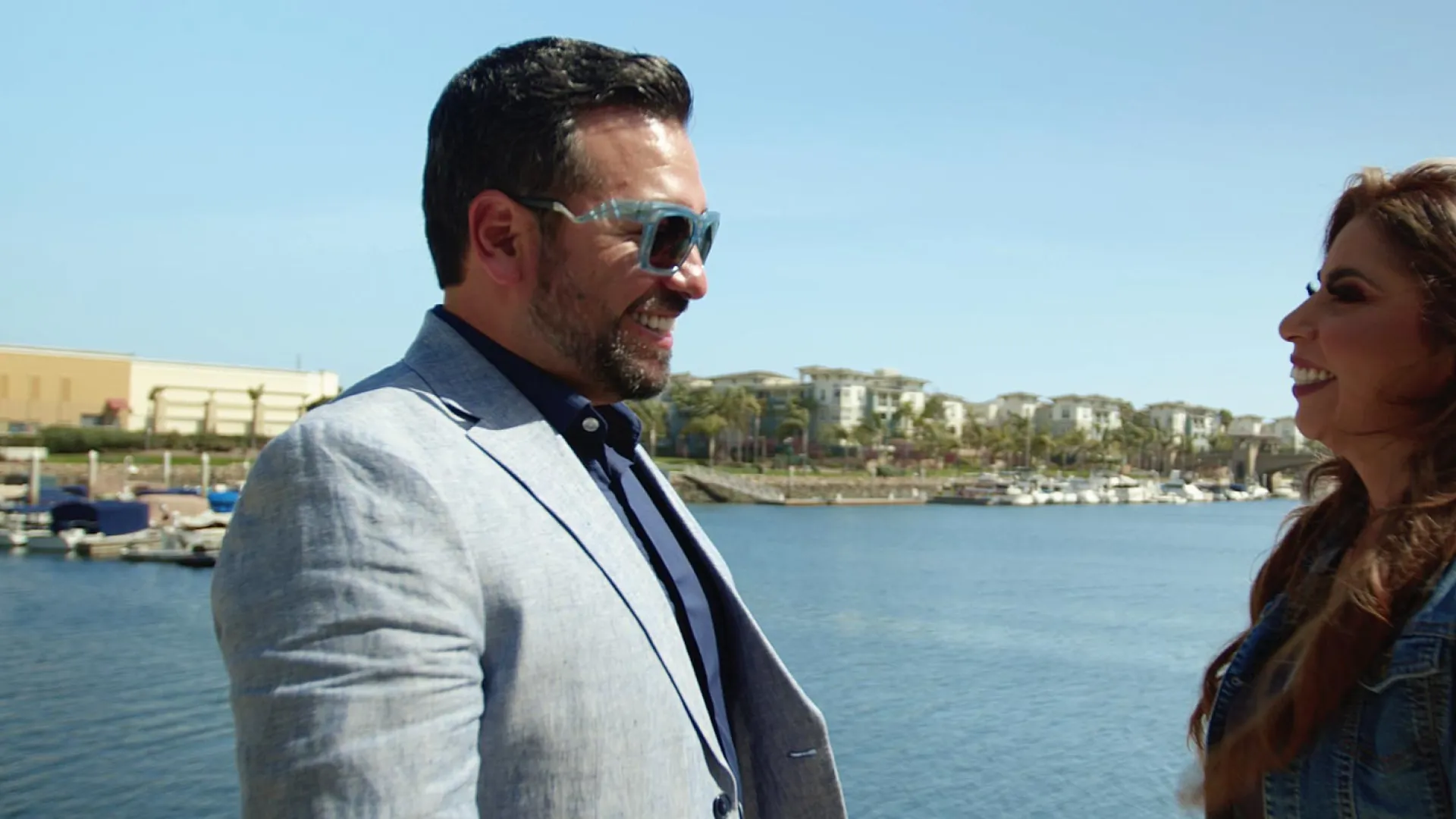 Two people smiling by the waterfront, boats and buildings in the background under a clear blue sky.