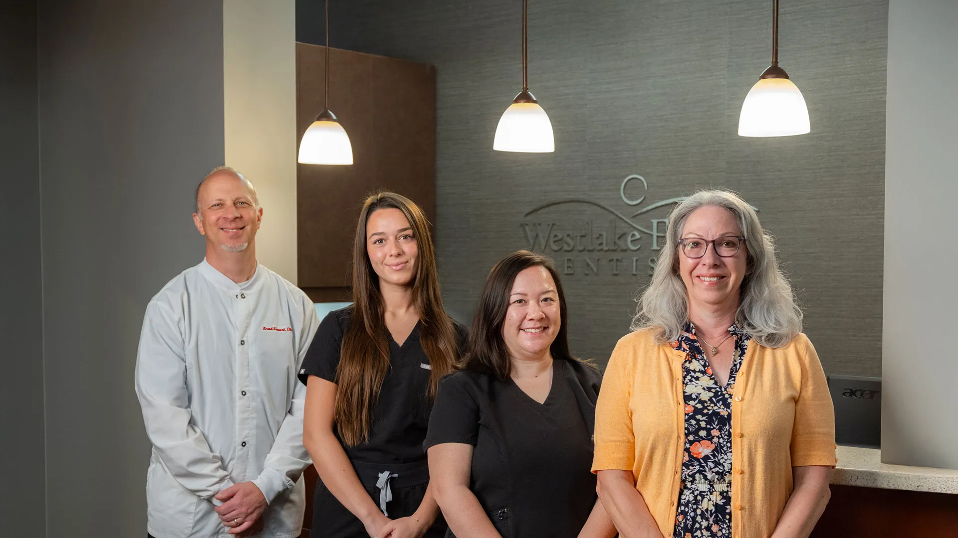 Four dental professionals smiling in a modern dental office with hanging lights and a reception desk.