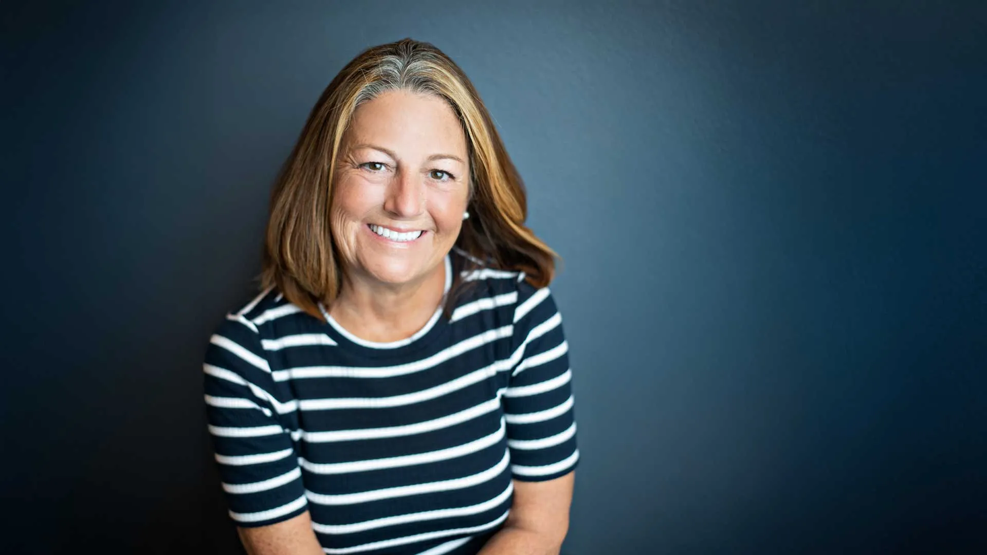 Smiling woman with shoulder-length blonde hair wearing a black and white striped shirt against dark background