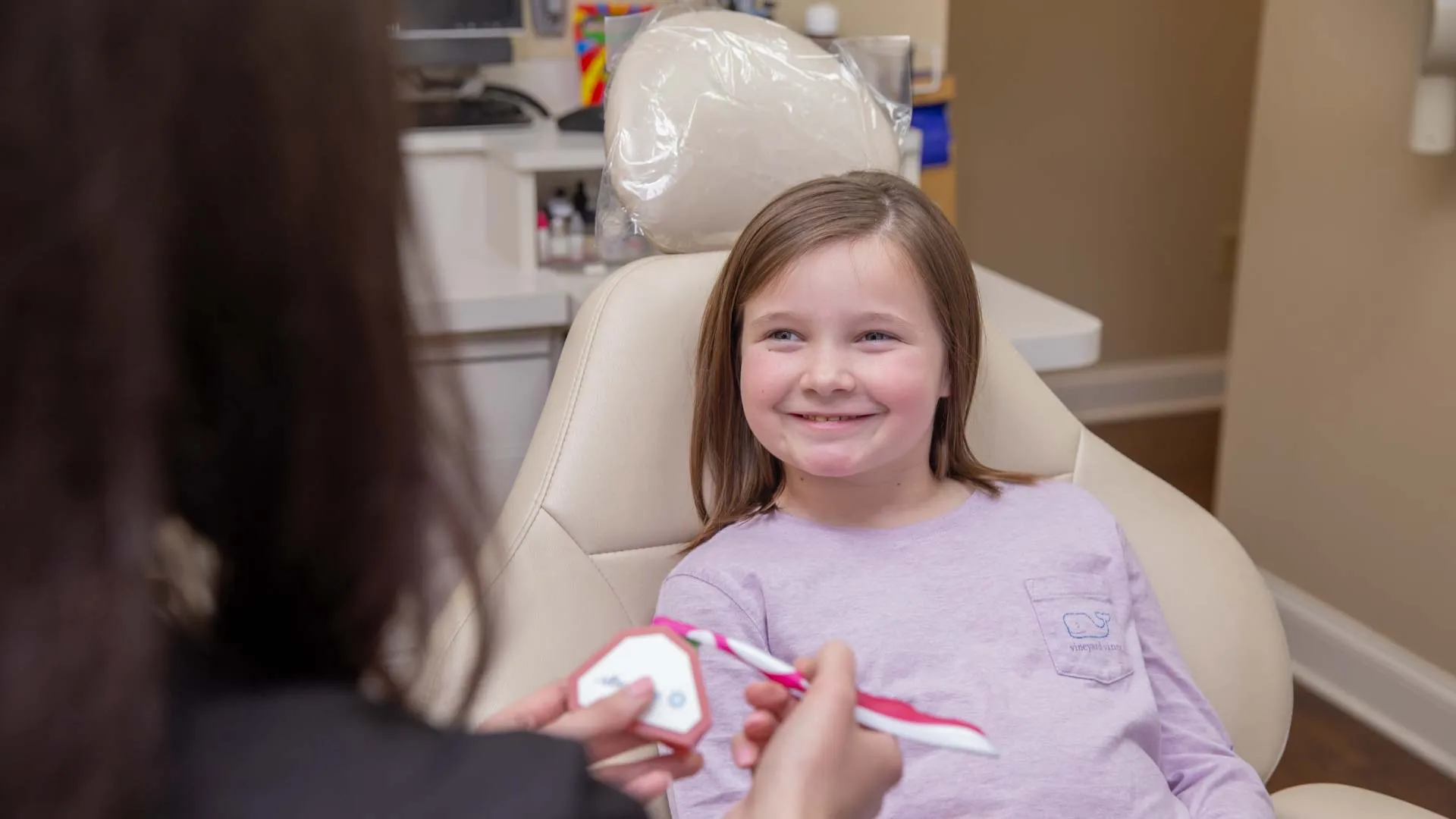 Smiling young girl in dental chair learning toothbrushing techniques from dental professional using model.