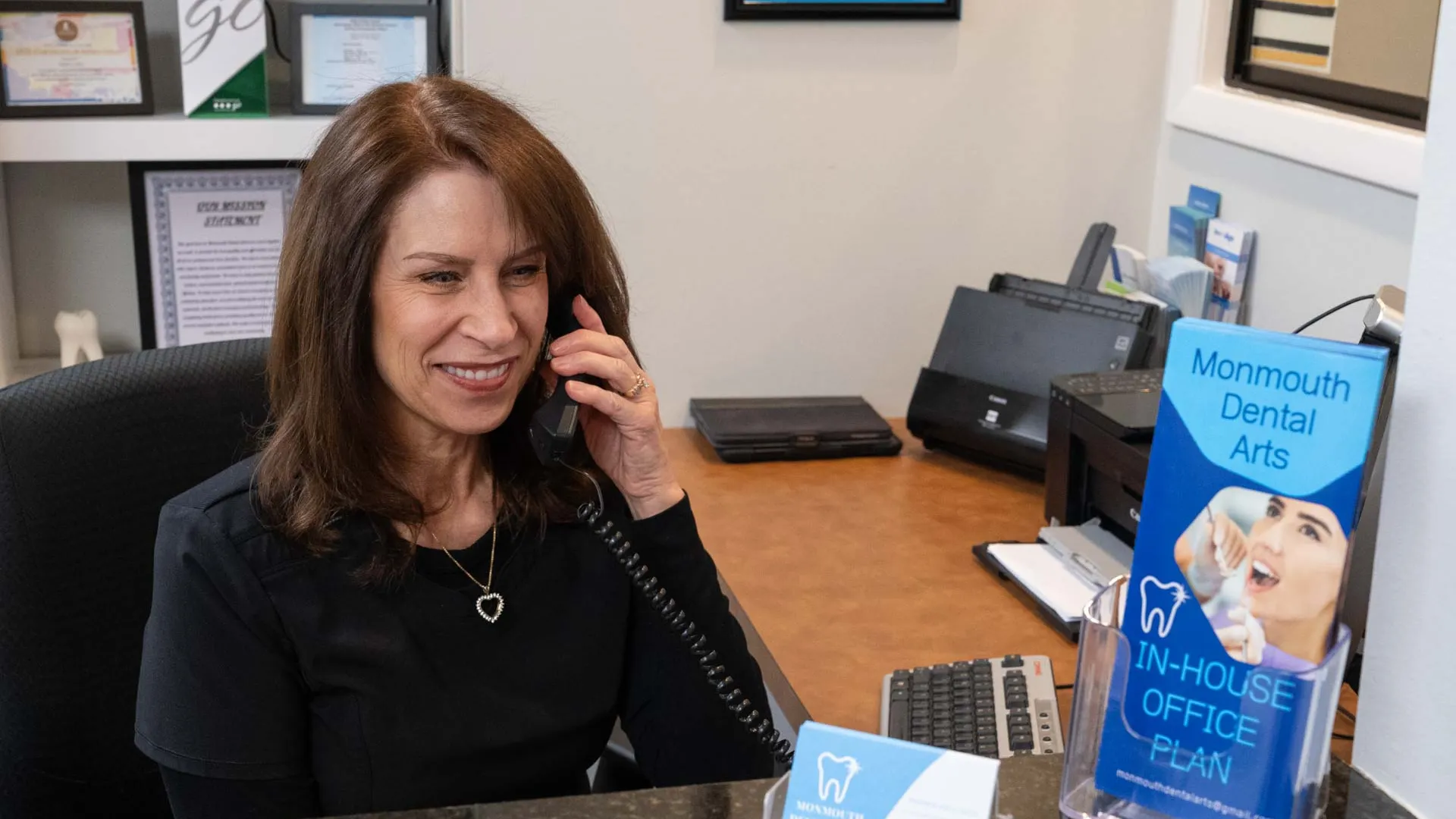 Receptionist smiling while speaking on the phone at a dental office with brochures displayed on the counter.