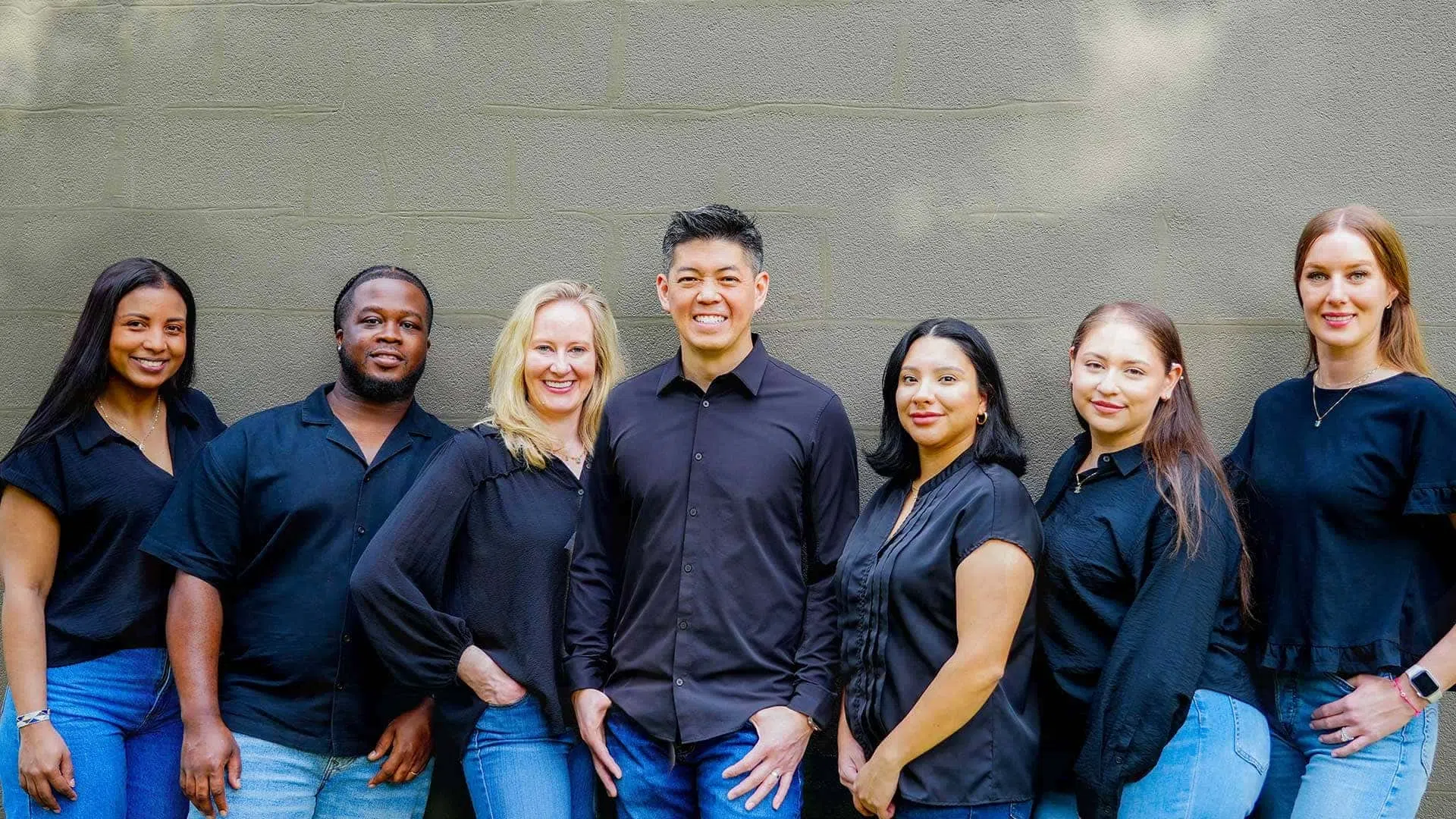 Diverse group of seven young professionals standing against a gray wall wearing black tops and blue jeans.