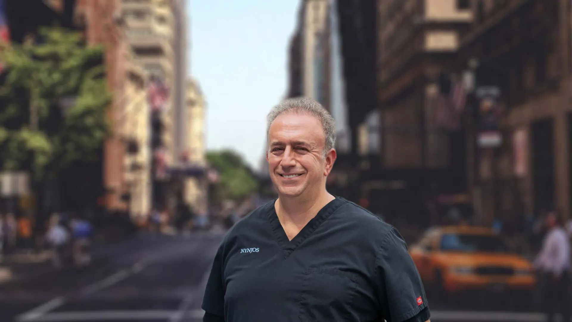 Smiling middle-aged man wearing black scrubs standing on a city street with blurred buildings and traffic in background