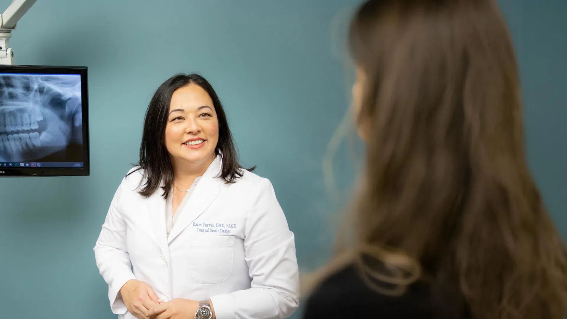 Female dentist in white coat smiling while talking to a patient with dental X-ray visible on screen