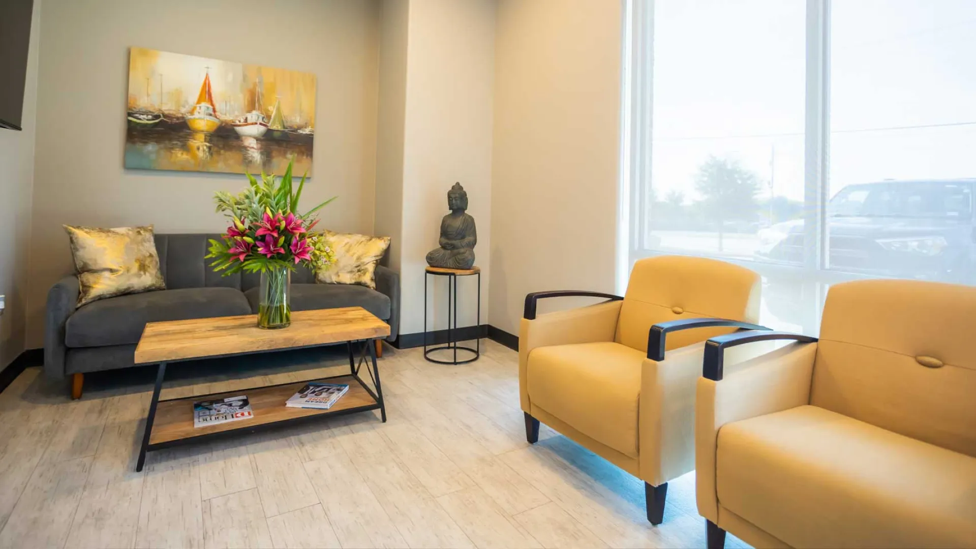 Modern waiting room with gray sofa, yellow chairs, wooden table with flowers, Buddha statue, and large window.