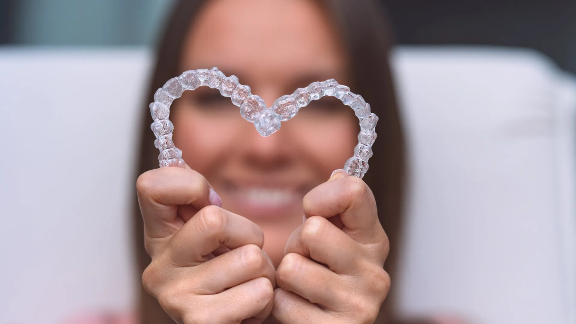 Smiling woman holding clear dental aligners shaped into a heart symbol close to the camera.