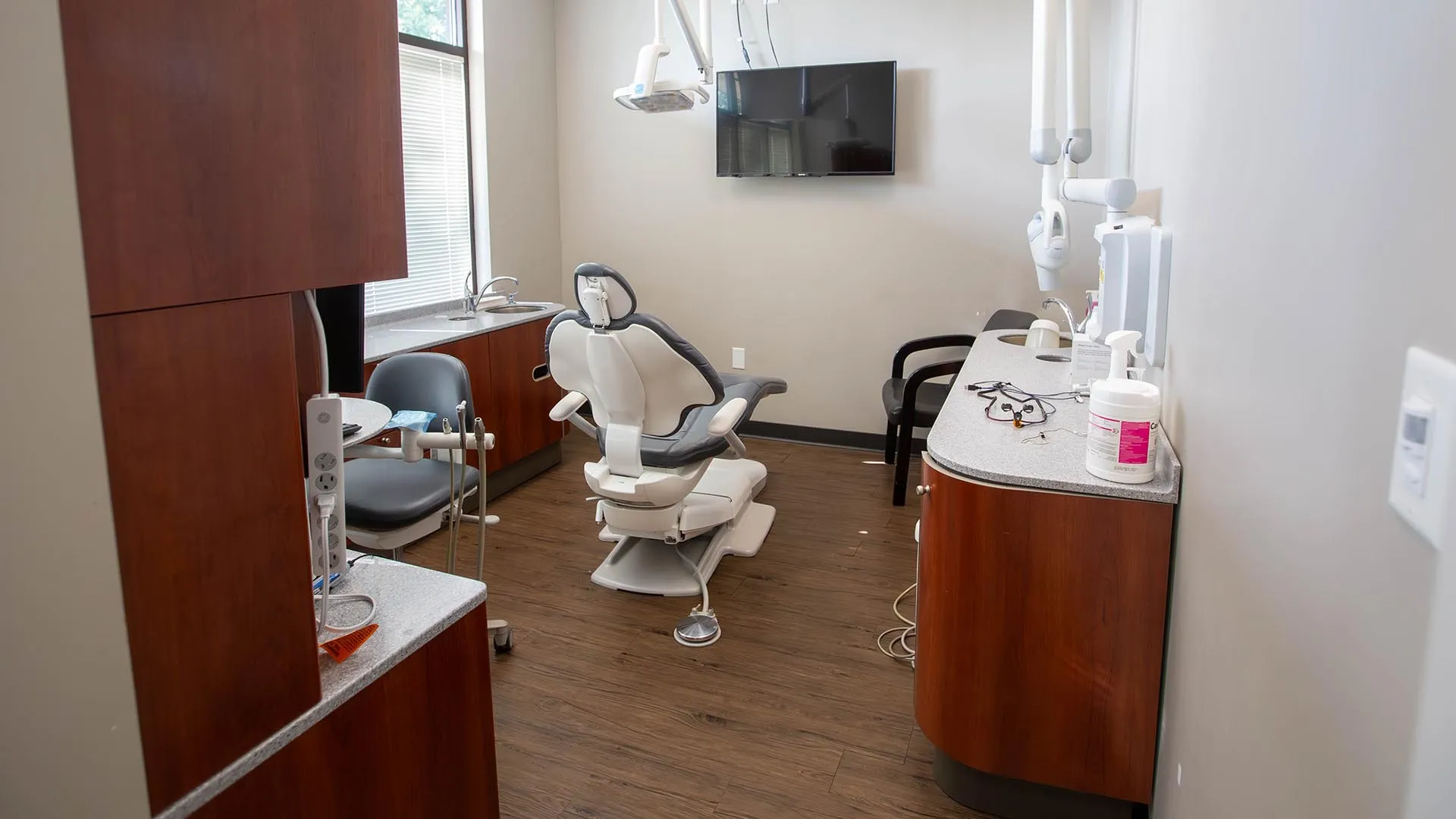 Modern dental office with patient chair, dental equipment, wooden cabinets, and wall-mounted TV screen.