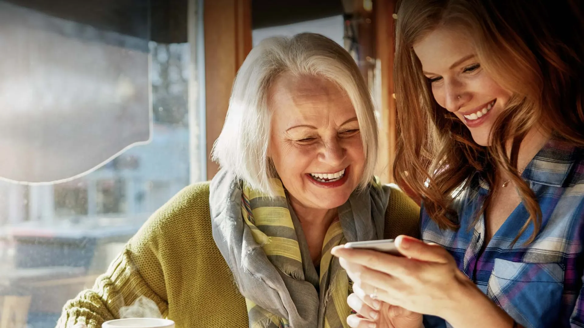 Two women enjoying coffee and sharing a moment together at a cozy café, smiling at a smartphone.