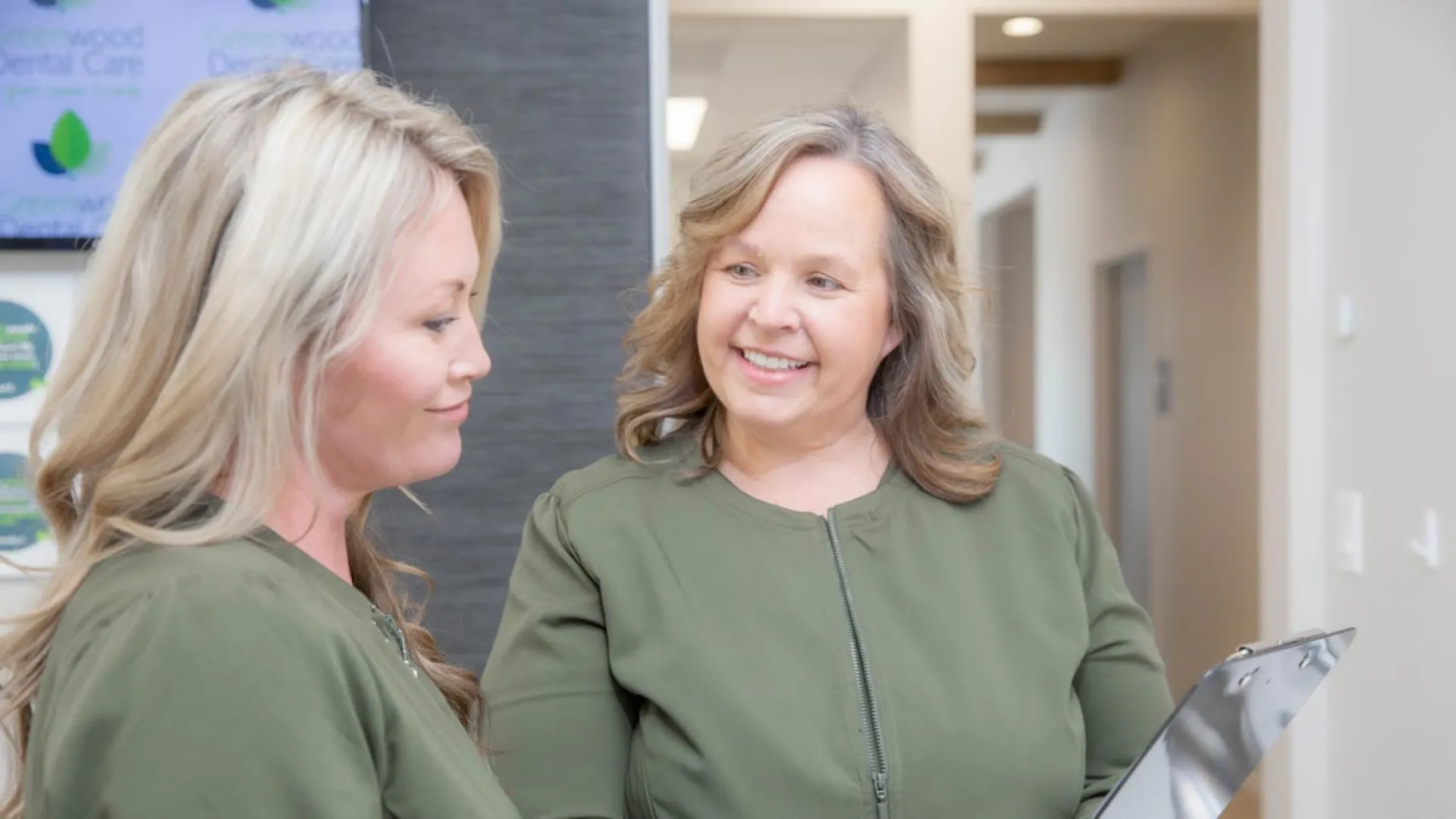 Two smiling women in matching green tops discuss a clipboard in a bright, modern office hallway.
