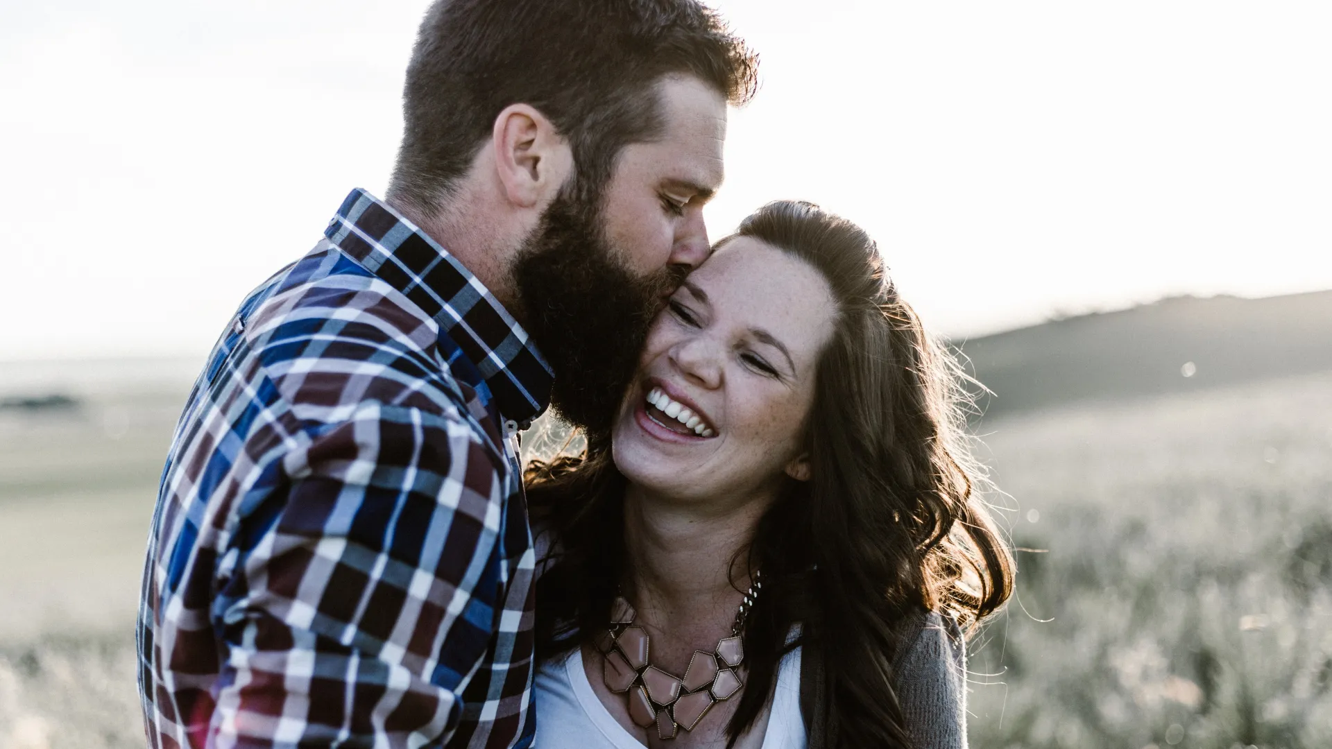Man in plaid shirt kissing smiling woman with necklace outdoors in a sunlit natural setting.