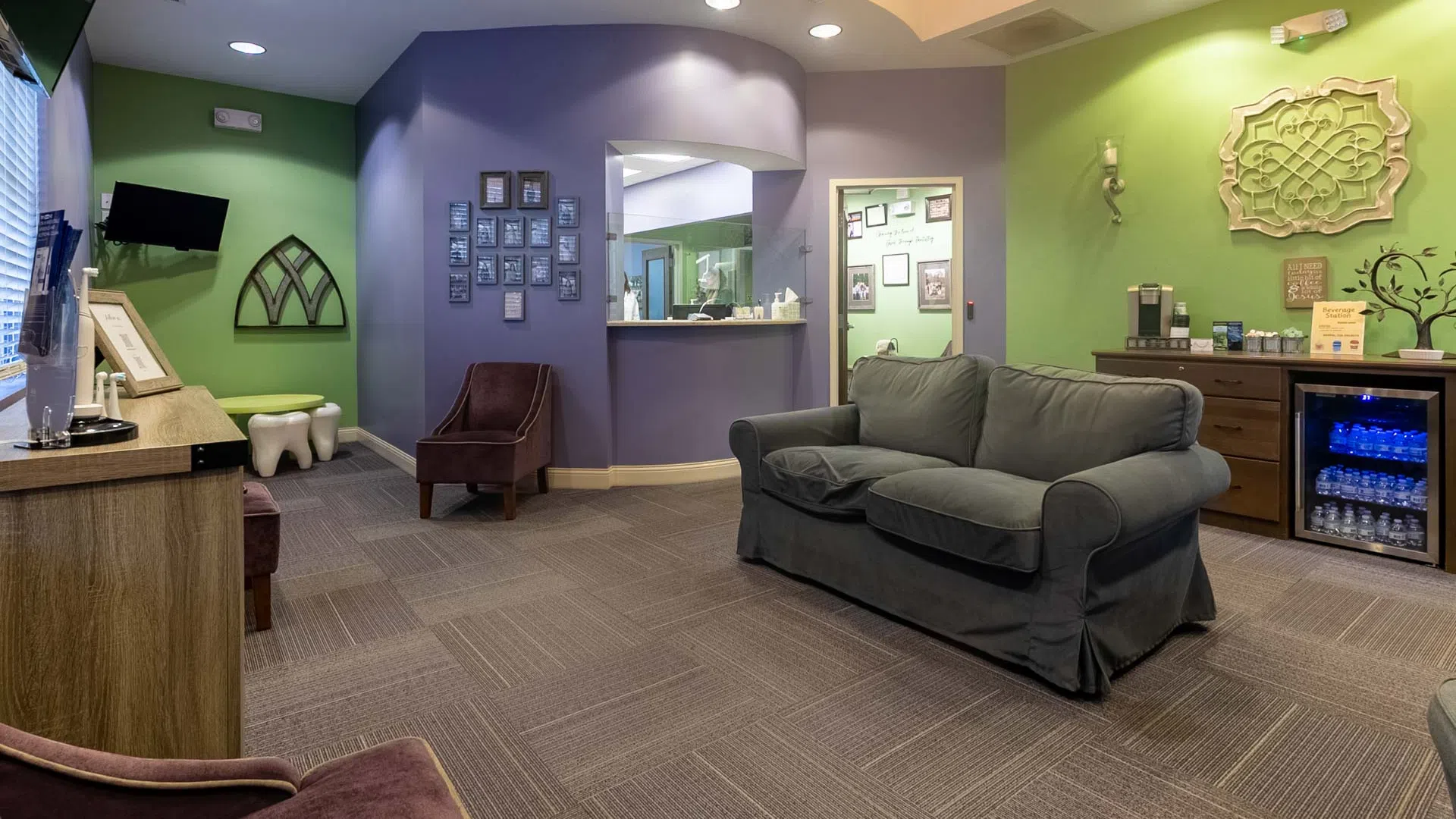 Cozy waiting room with gray sofa, purple and green walls, TV, chairs, and a refreshment station with water and coffee.