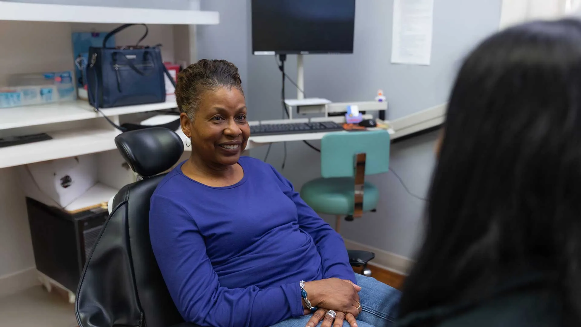 Smiling woman in blue shirt sitting in chair, talking to someone in an office with computer and shelves.