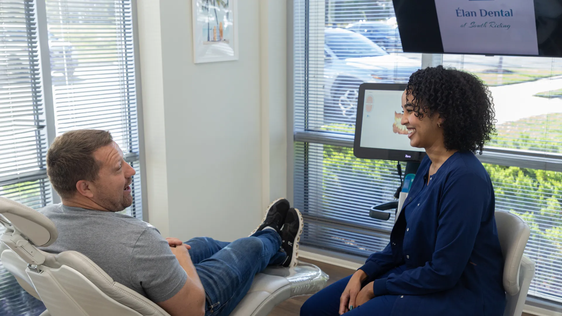 Dentist and patient smiling and discussing treatment in a bright dental clinic with large windows.