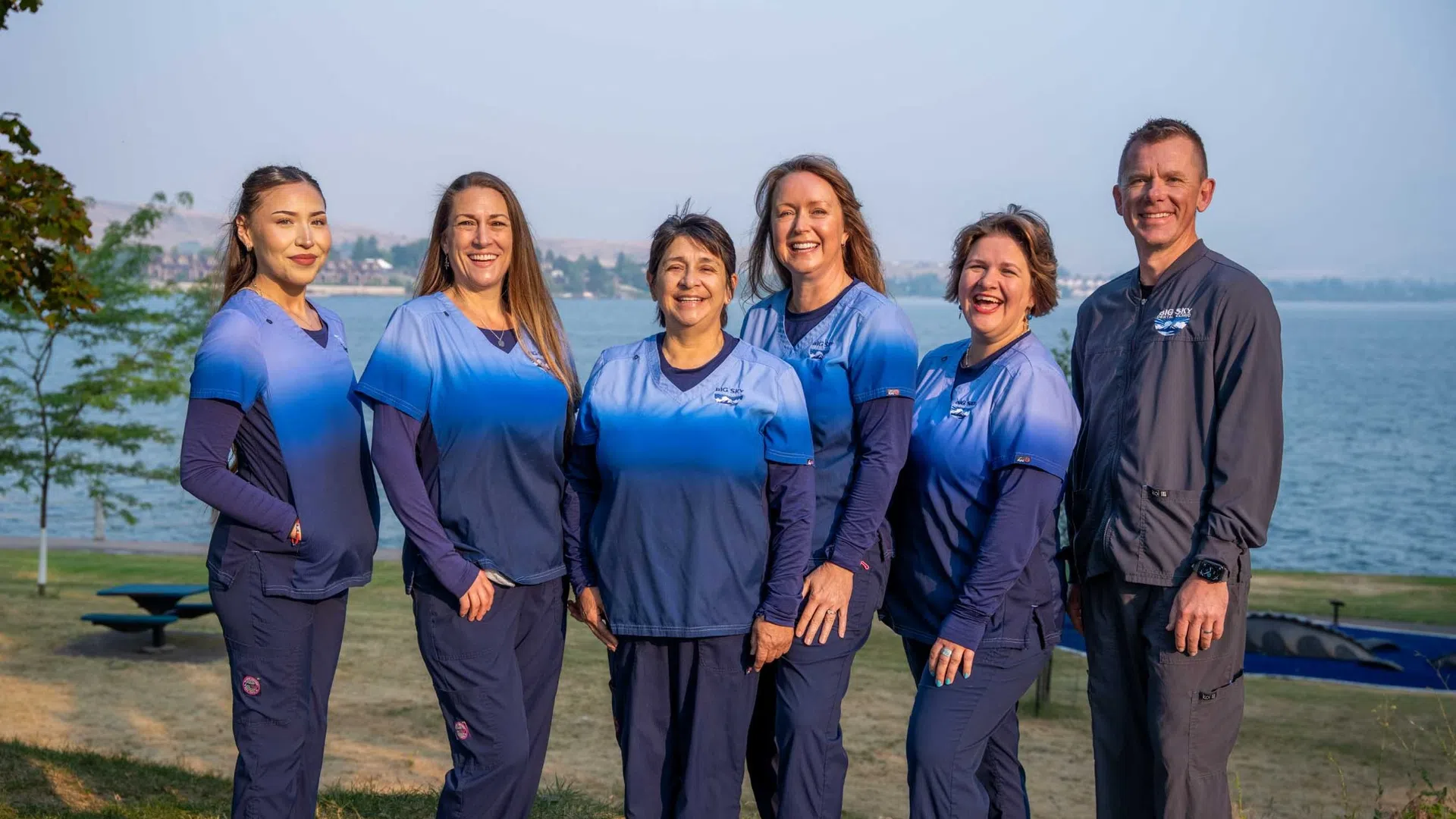 Group of six medical professionals in blue scrubs standing outdoors by a lake on a sunny day