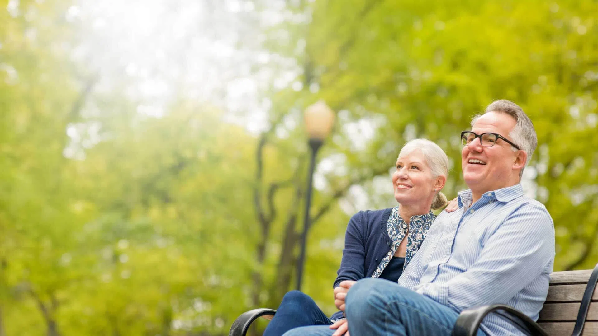 Happy senior couple sitting on a park bench enjoying a sunny day surrounded by green trees and nature.